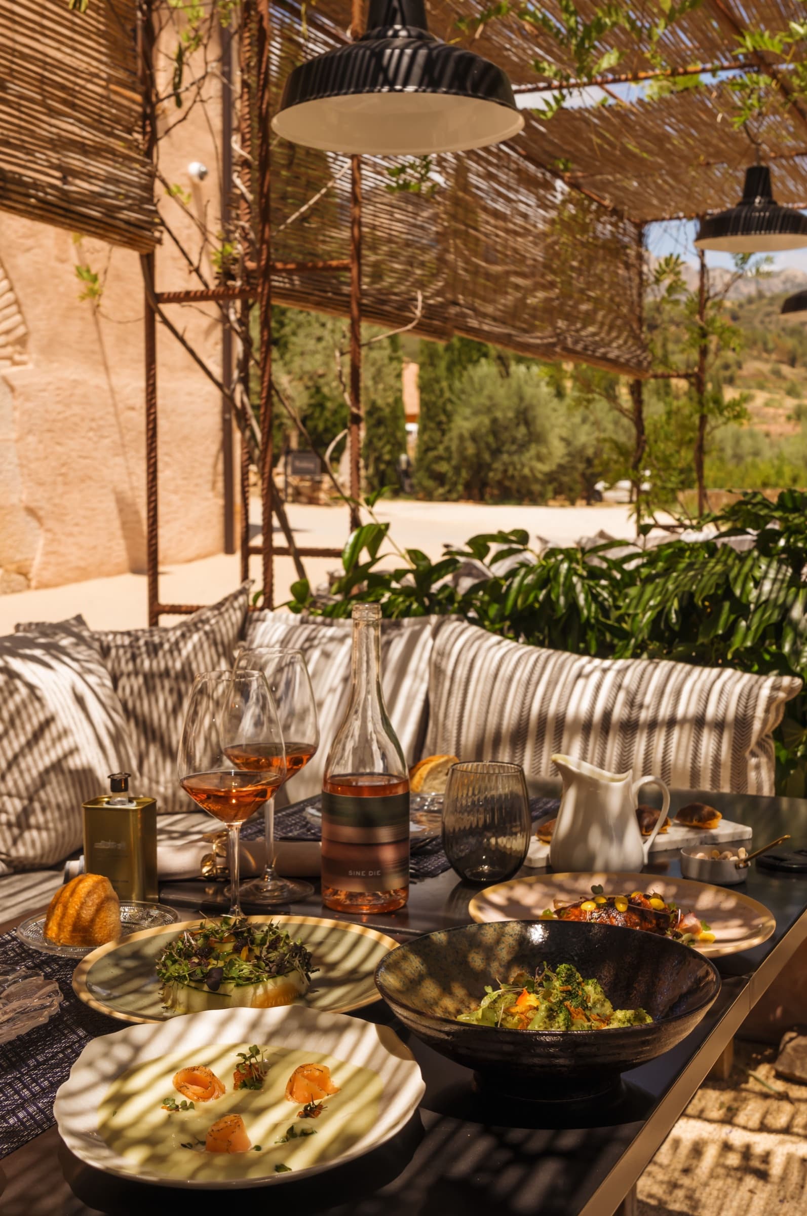 A summer lunch on the outdoor pergola — striped linen pillows, plates of vegetables on dark stoneware, a bottle of rosé, a glass carafe of olive oil, dappled light through reed shade overhead.