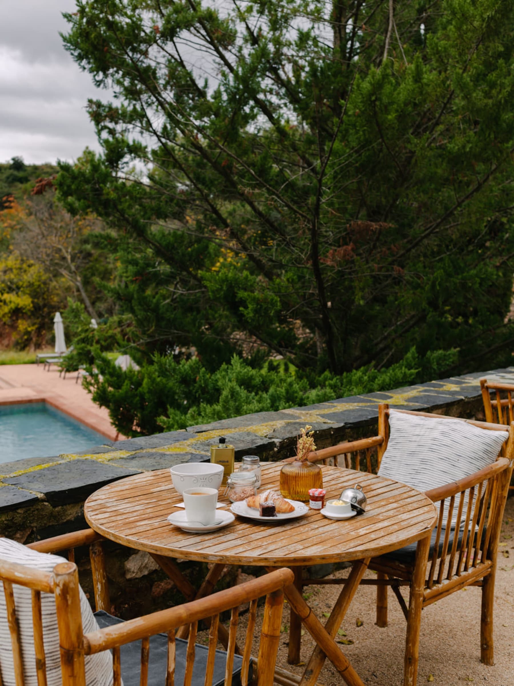 A small round teak breakfast table for two beside the pool — a cup of coffee, a croissant, a small jar of jam, a pine tree leaning over from the bank above.