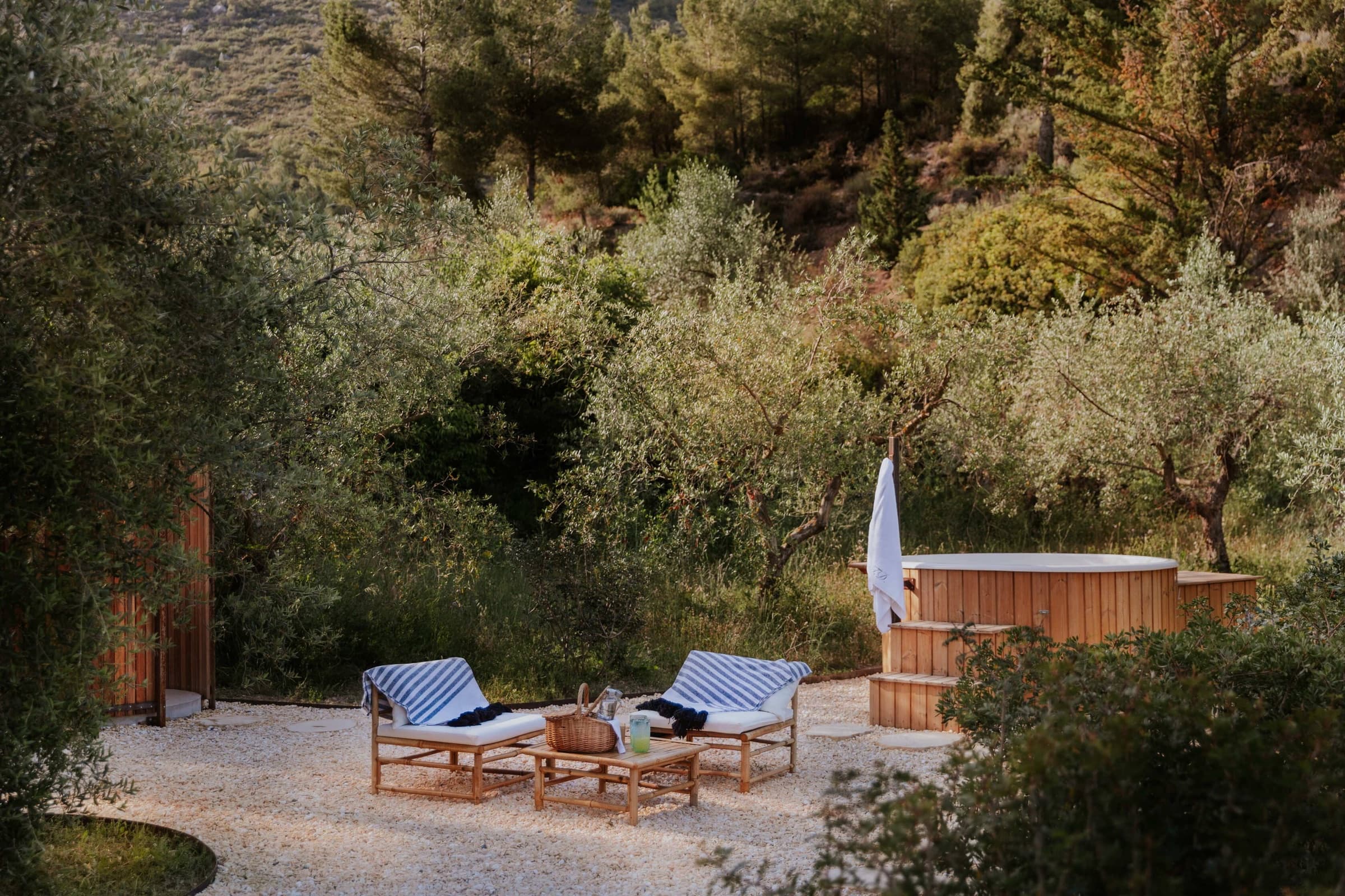 An outdoor cedar soaking tub on a gravel platform, two striped sun loungers and a low table beside it, olive trees screening the slope behind.