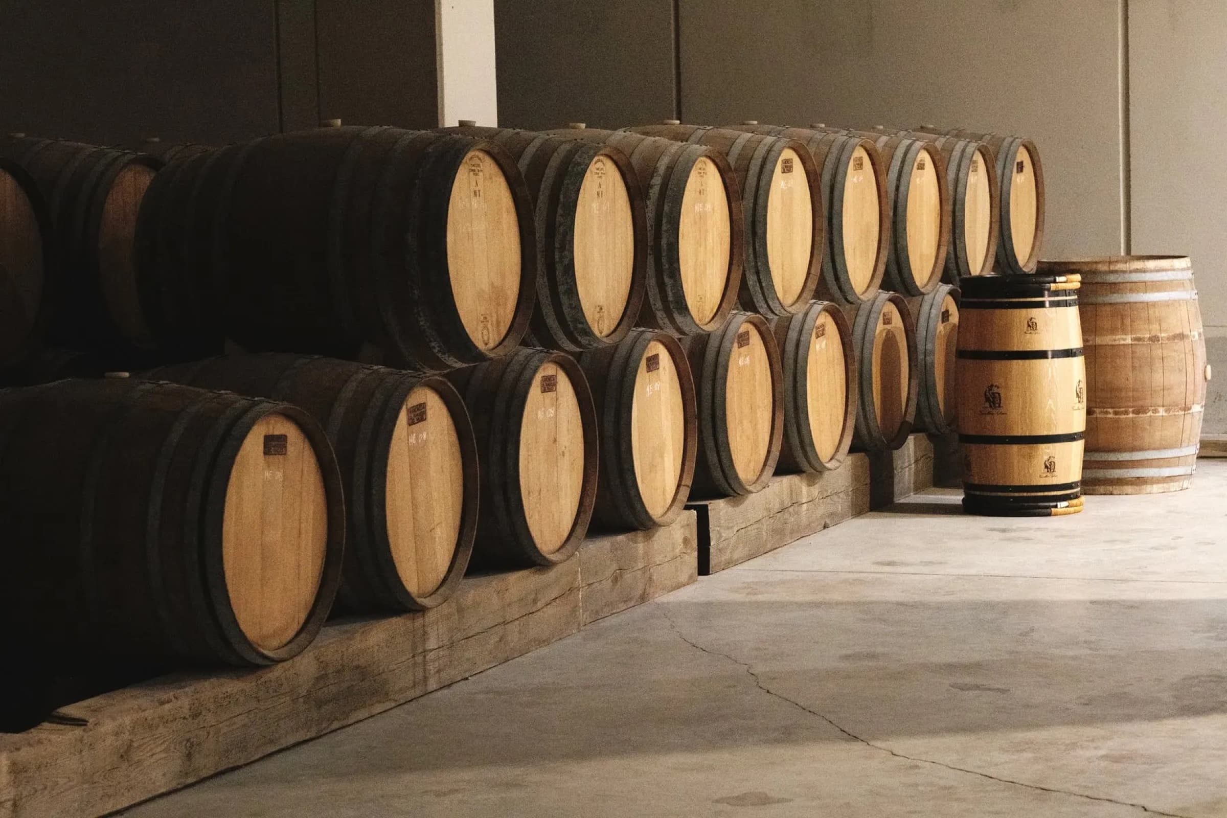 Stacked rows of oak barrels in the on-site cellar, the lighter heads of the upper row in soft warm light against the darker bodies below.