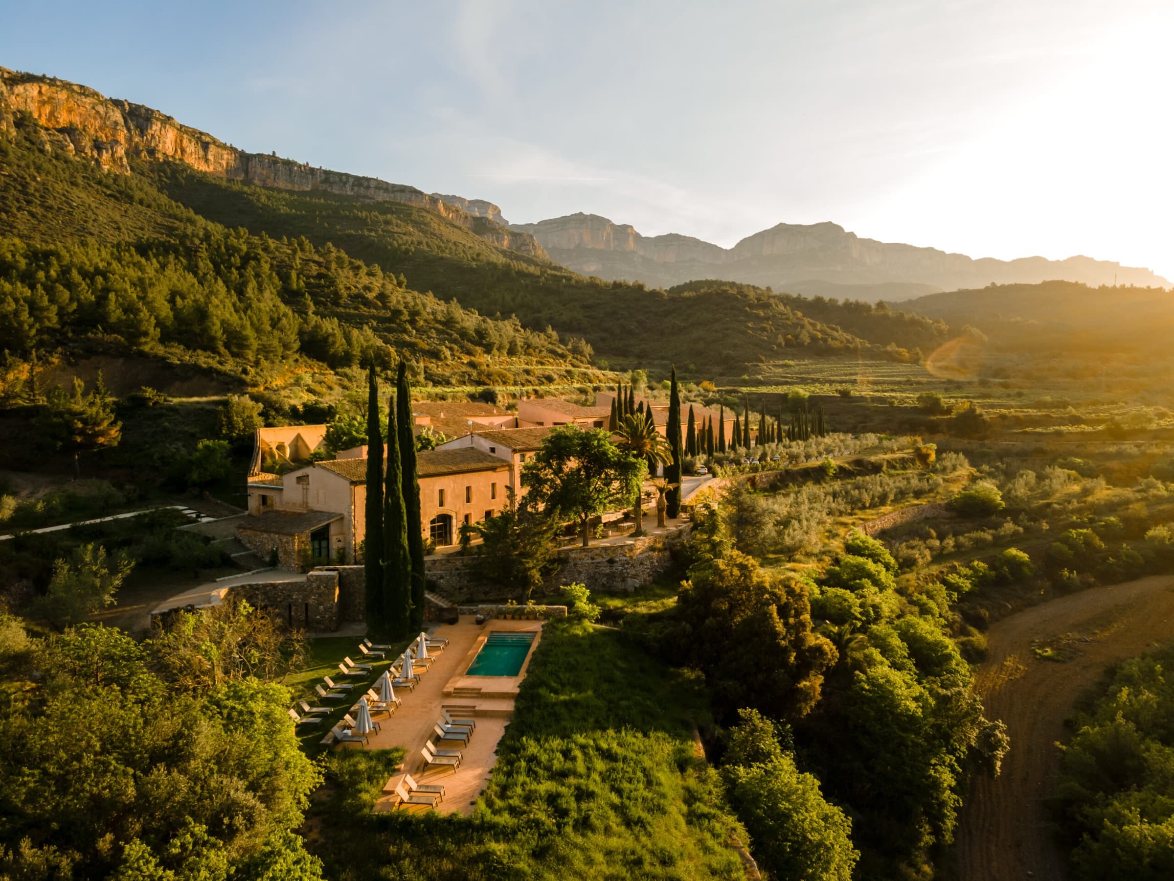An aerial view of the restored stone hamlet at golden hour — tall cypresses around the buildings, a long lap pool on the lower terrace, olive groves and vineyards descending to the valley, the Serra de Montsant ridge beyond.