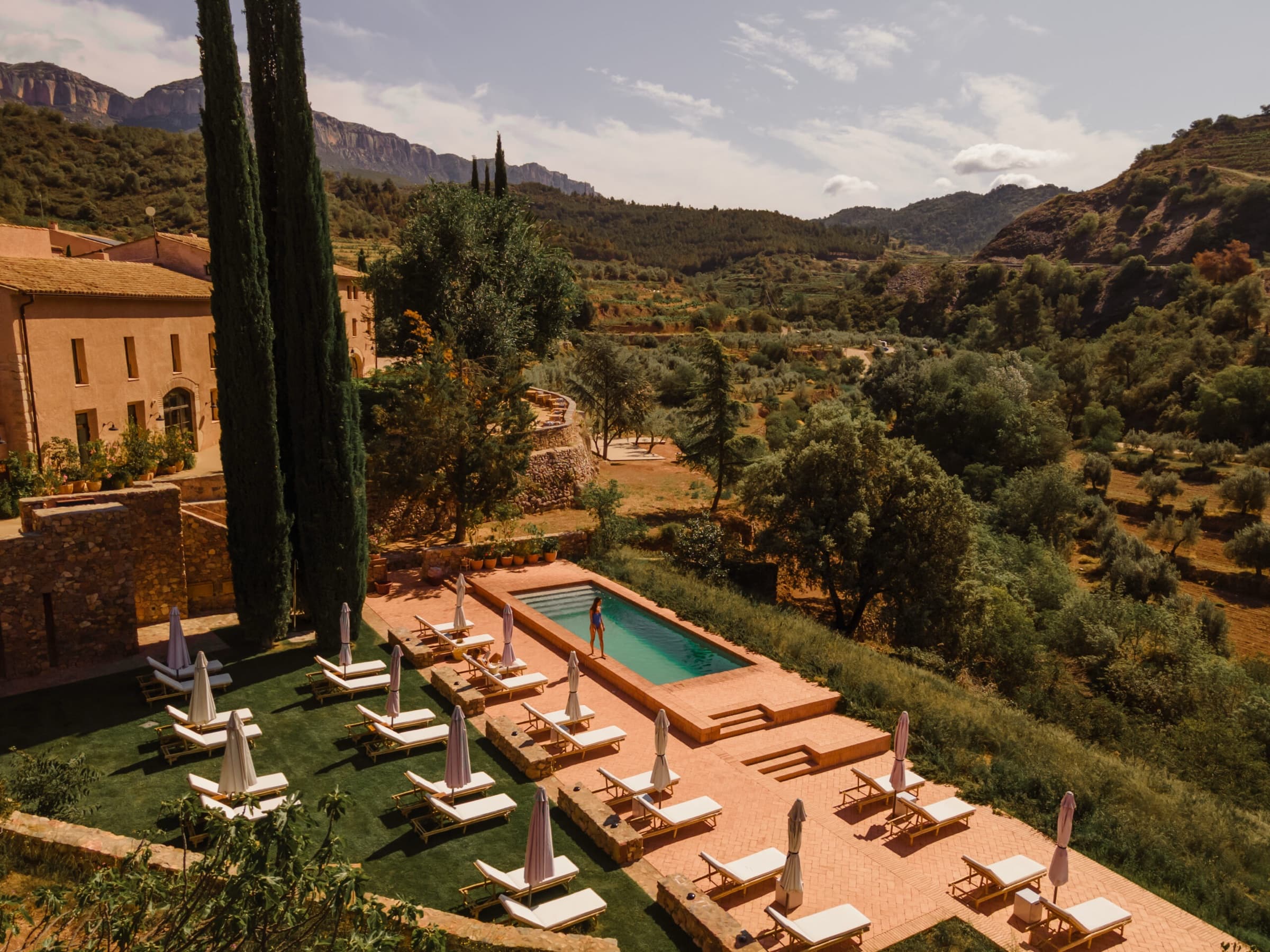 A long pool on a slate terrace above terraced olive groves, sun loungers along the side, the wooded slopes and grey limestone ridge of the Serra de Montsant in the distance.