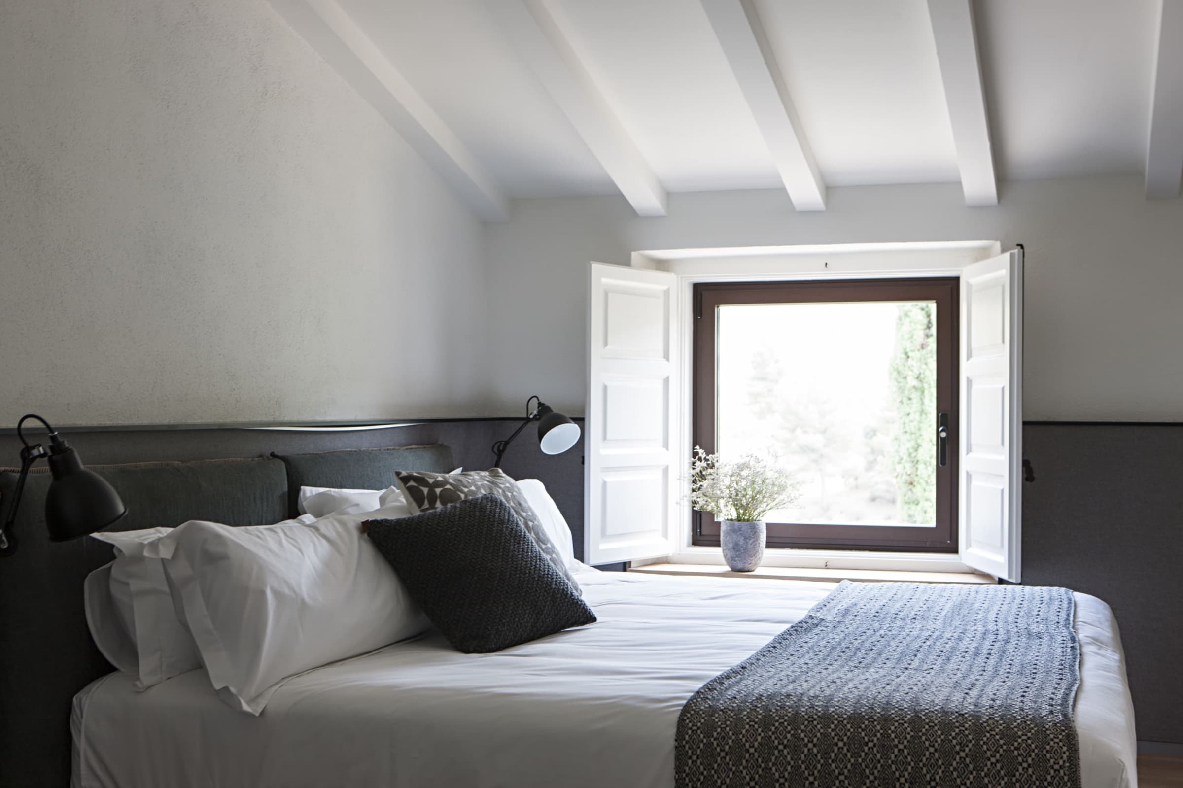 A guest room with a pitched white-beamed ceiling, charcoal headboard and wainscot, white linen, and an open casement window onto cypresses and the morning hills.