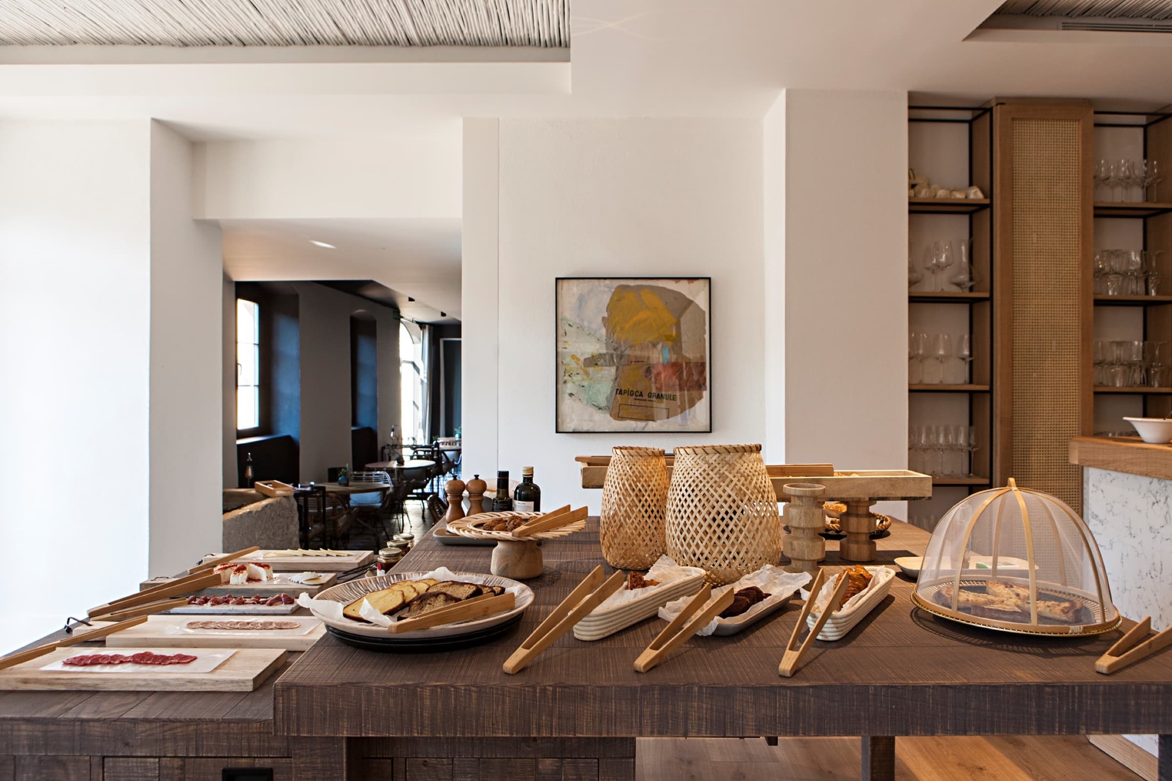 A long oak breakfast buffet — boards of charcuterie and cheese, woven covers over warm pastries, bottles of olive oil, glassware on caned shelves behind, a small framed painting on the white wall above.