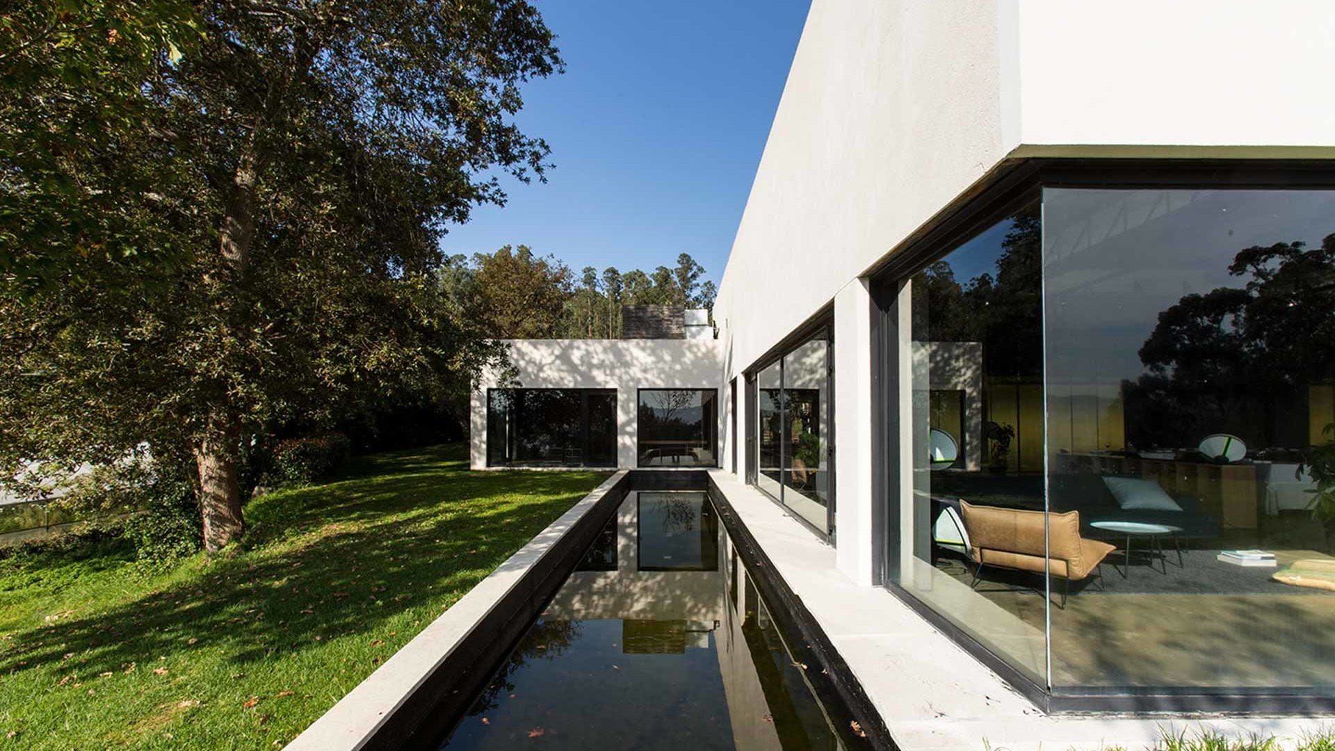 The pavilion seen by daylight from the side, a long reflecting pool below the glazing and the Galician woodland behind.
