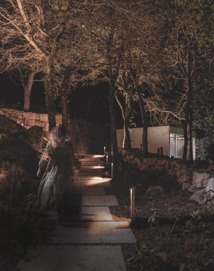 A guest in a long dress walking a stone path lit at intervals through the woods toward a lit galpón.