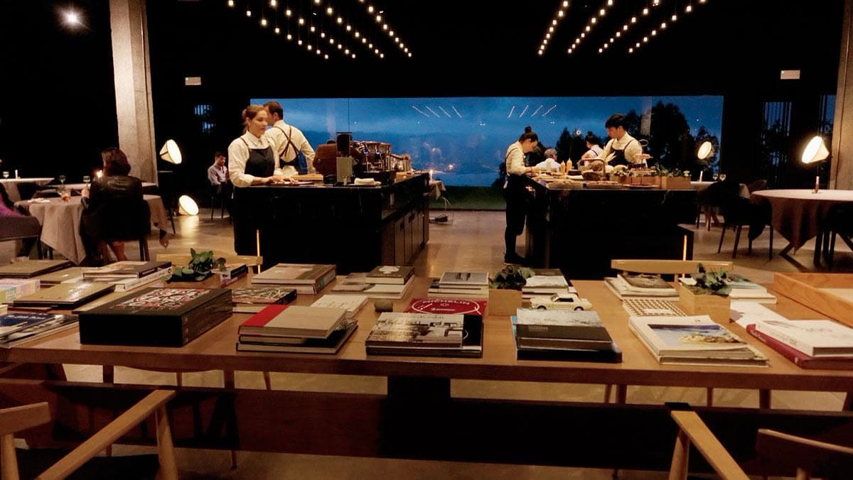 The dining room with the open kitchen behind, low festoon lights and an evening service set against the long western window.