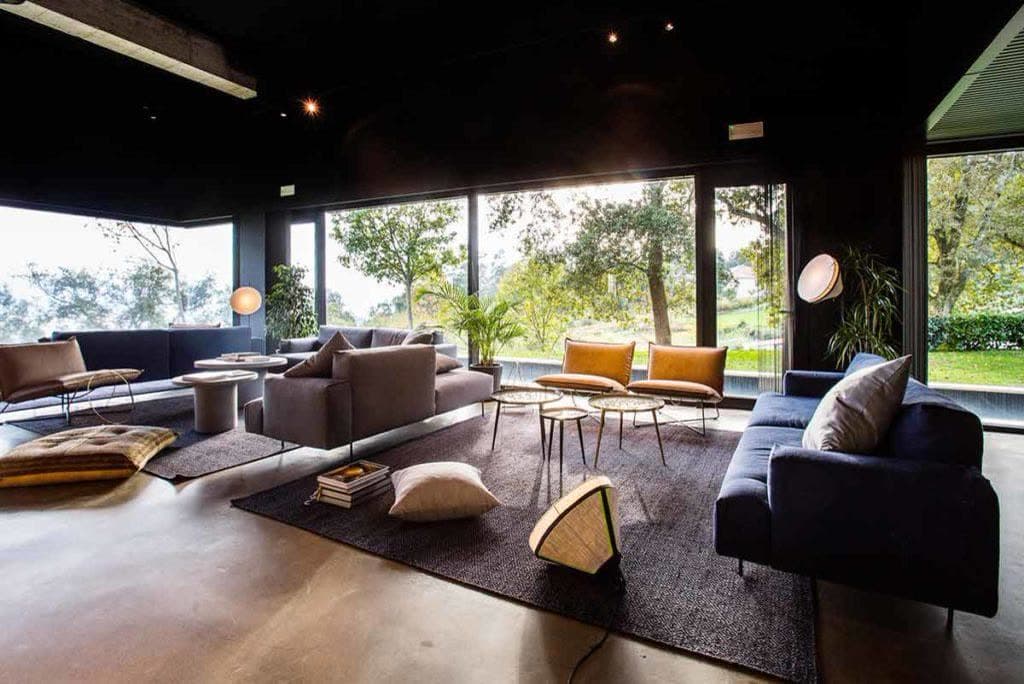 The lounge — low chairs and a tan leather lounger in front of full-height glazing onto a clearing of tall trees.