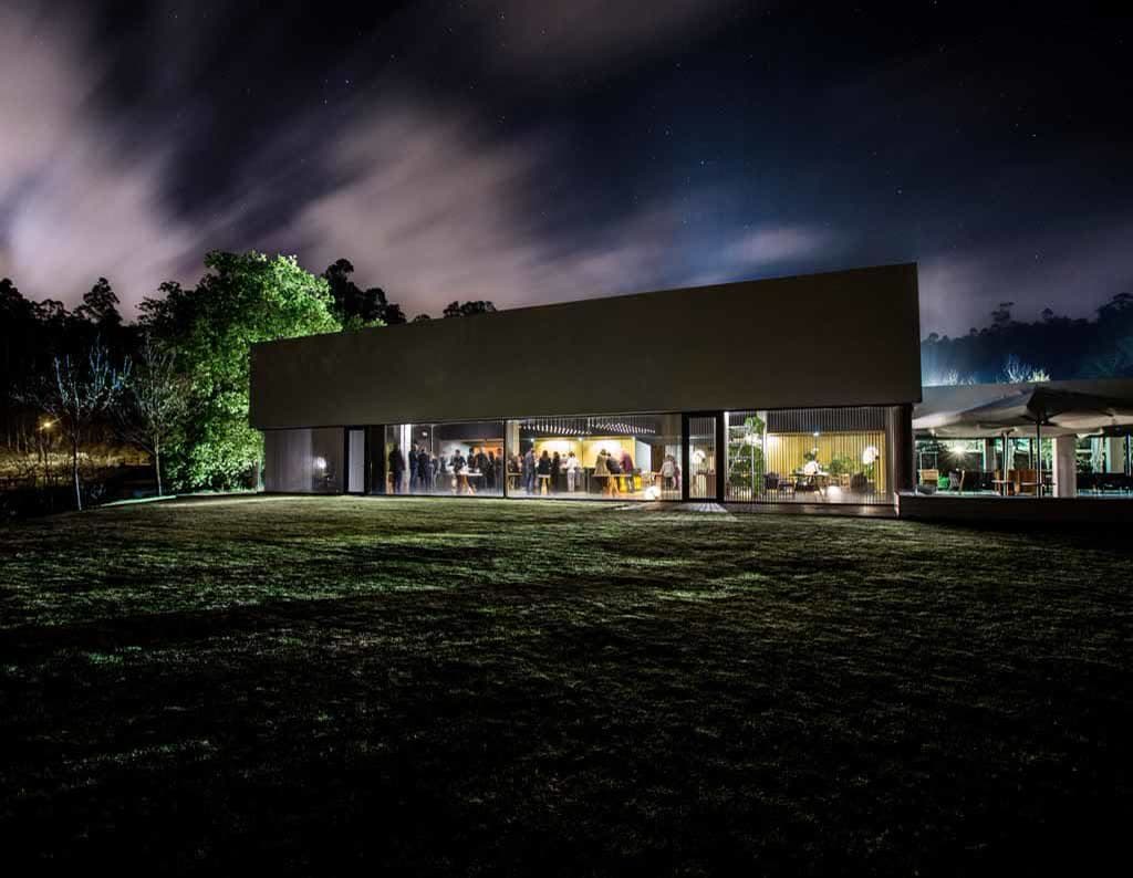 The Pepe Vieira pavilion seen at night under a darkening sky, the dining room glowing through tall glazing onto a lawn at the edge of the woods.