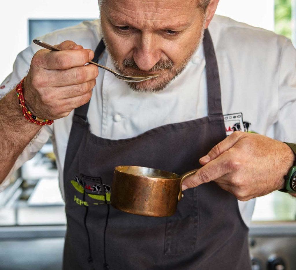The chef tasting from a small copper pan in close view, grey apron and a quiet hand at the centre of the kitchen.