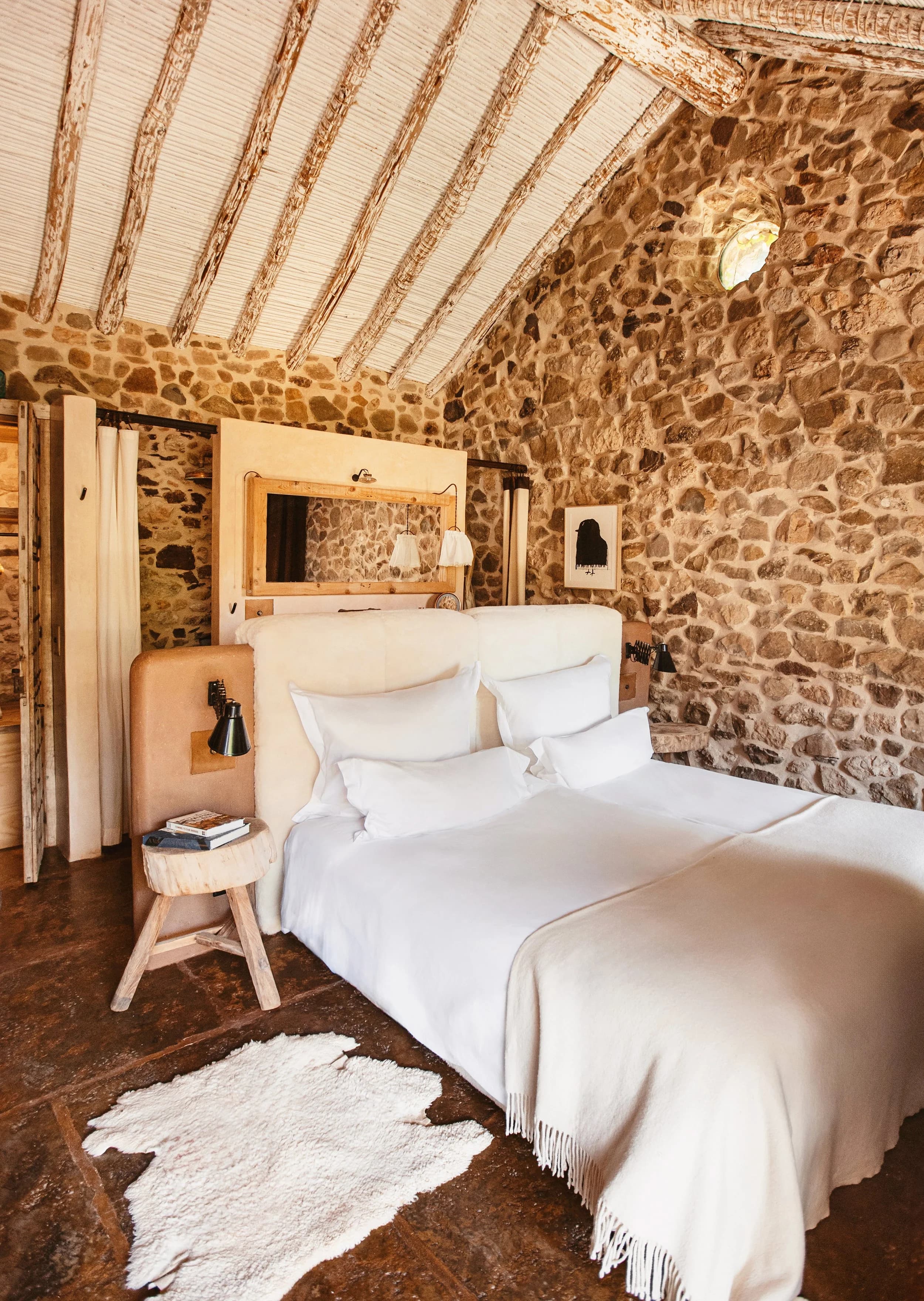 A bedroom inside the cortijo — pale dry-stone walls, white linen on a wide bed, exposed timber beams under a reed-and-lime ceiling.
