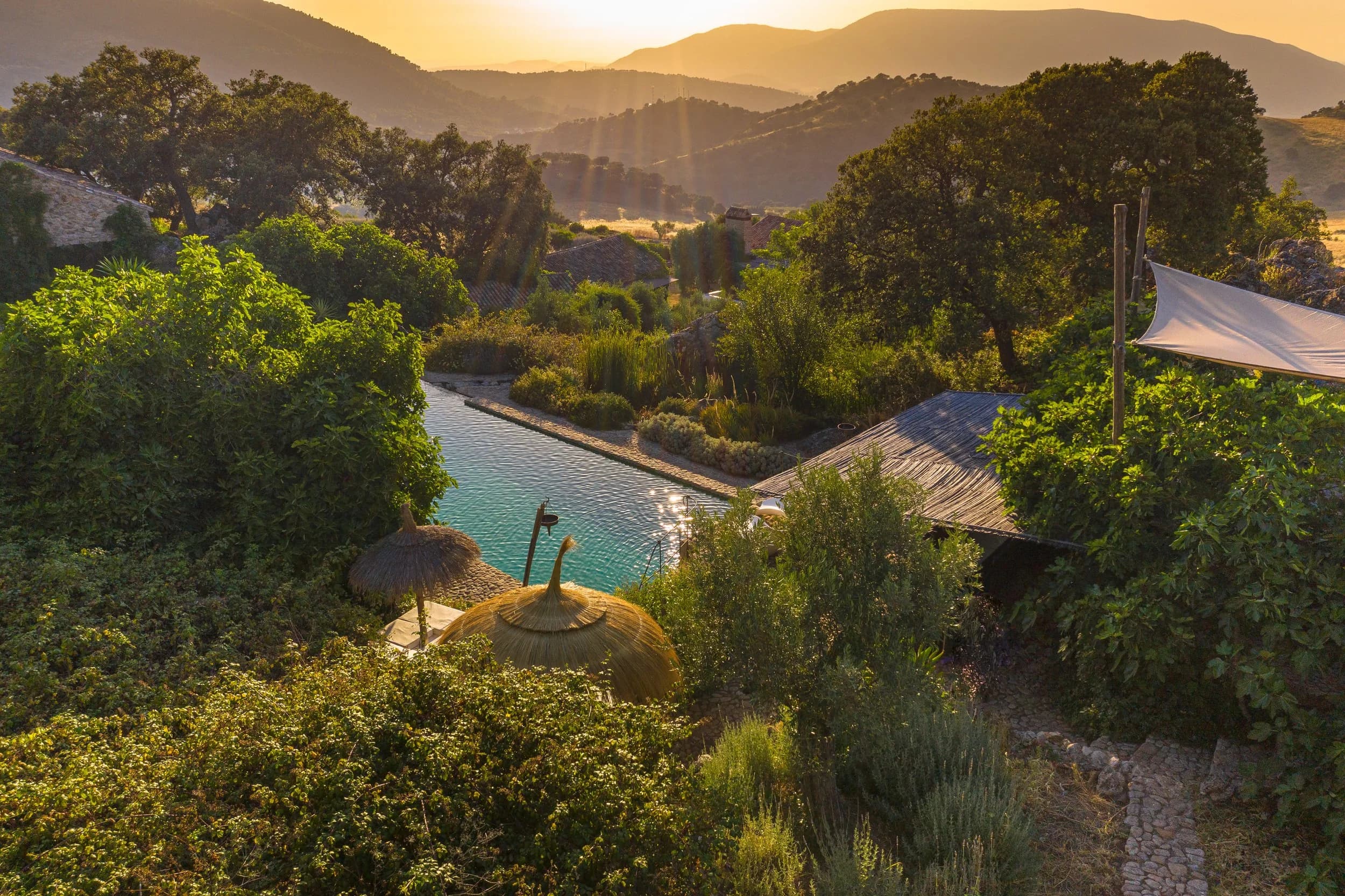 A natural stone-edged pool below the Andalusian sierra at dusk, soft tents and wild planting around it, the mountains opening toward the sun.