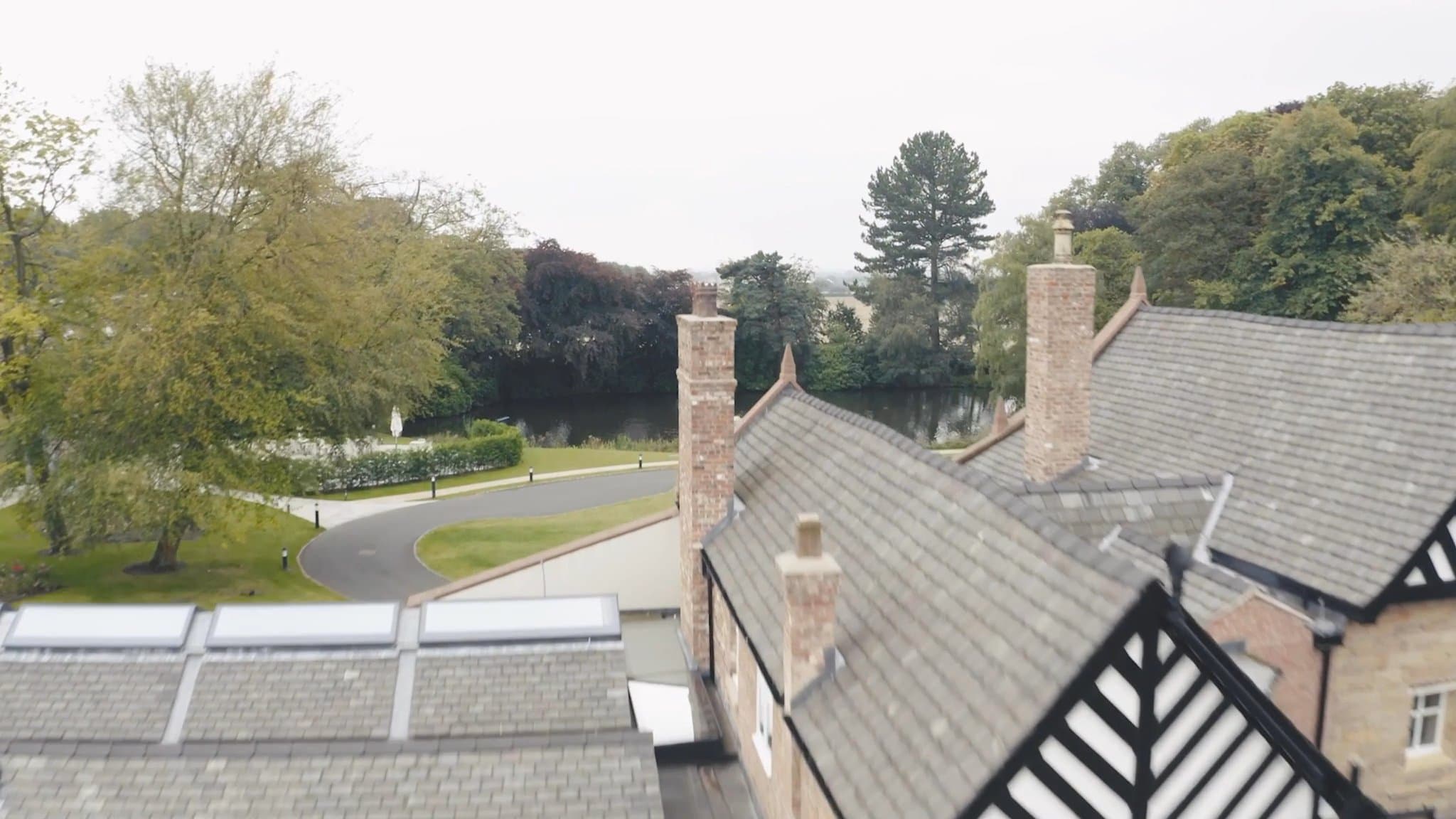 An aerial view across Moor Hall's tiled and slate roofs — three brick chimneys and a black-and-white timbered gable in the foreground, a manicured drive and the still lake beyond, framed by mature trees in early autumn.