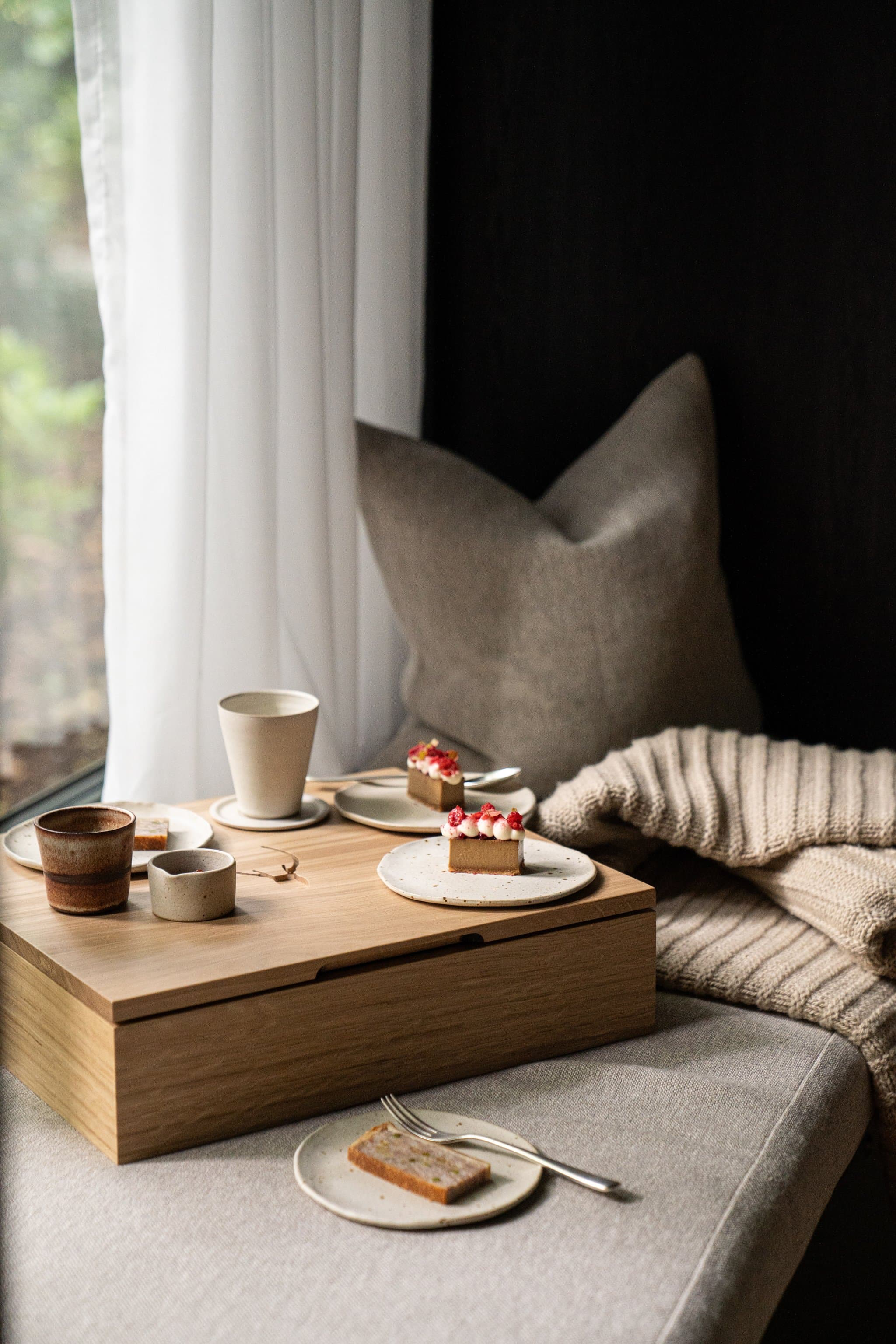 A small oak tray on a window-seat at Moor Hall — a pale ceramic mug of coffee, two espresso cups, and three thin slices of teacake topped with pink rhubarb on speckled stoneware plates, a cream knit throw and linen cushion folded behind.