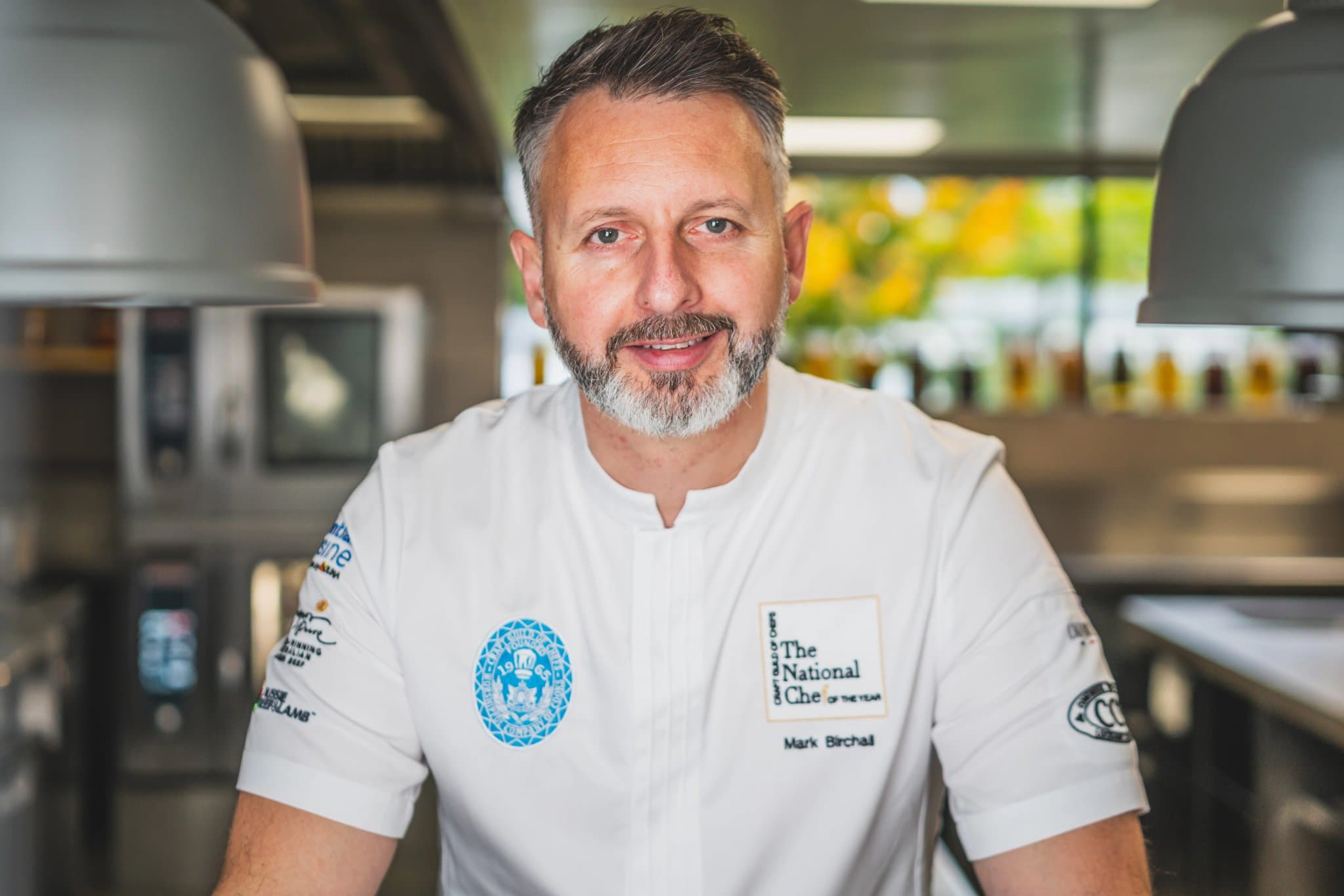 Mark Birchall in a white short-sleeved chef's jacket embroidered with the National Chef of the Year crest at the chest, standing at the kitchen line under two grey heat lamps, the polished steel pass and a soft kitchen-garden window behind him.