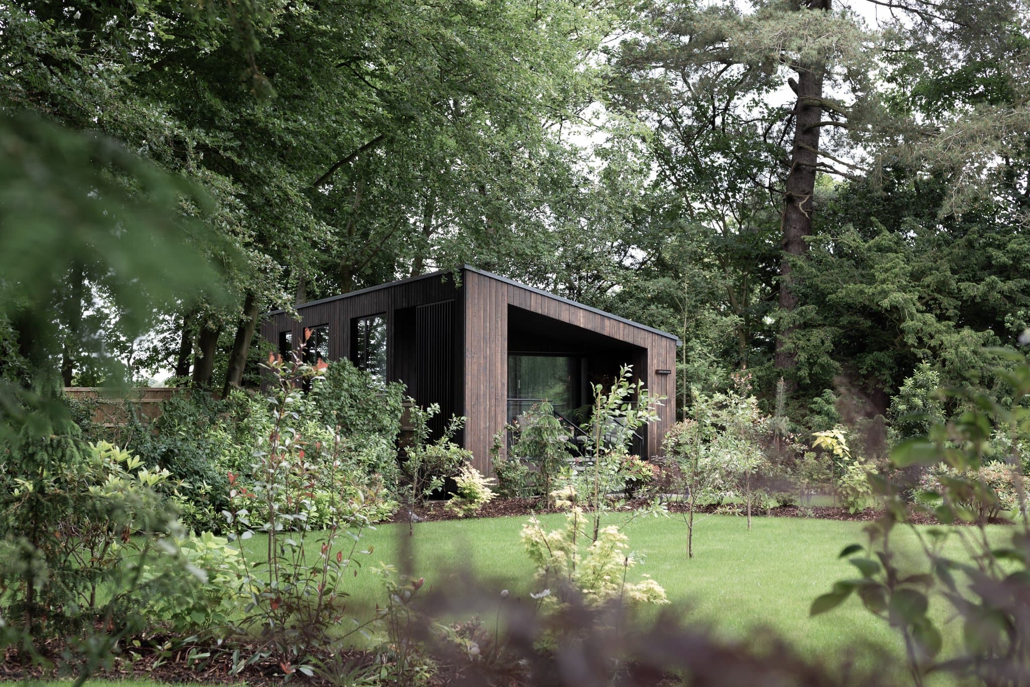 A black-stained timber-clad Garden Room set in long grass and trees on the Moor Hall meadow — a single low cabin with a tall picture window and dark trim, framed by birch, holly and red-leaved shrubs in the foreground.