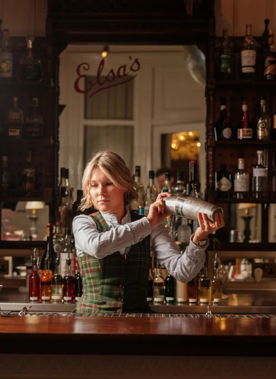 A bartender shaking a cocktail behind the polished bar at Elsa's, the room's name in cursive on the mirror behind.