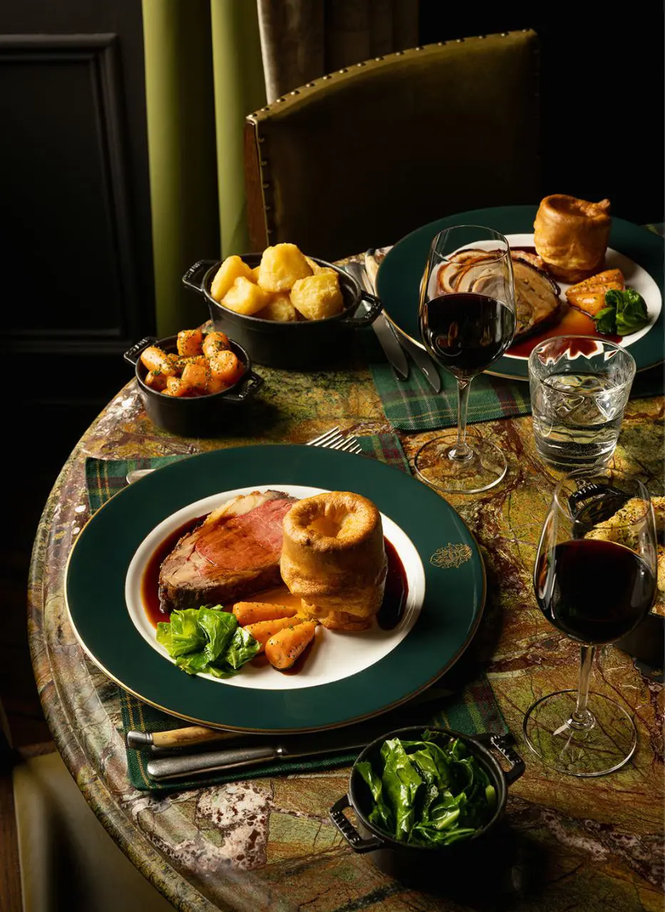 A Sunday roast laid at The Clunie — pink beef, Yorkshire pudding, greens and red wine on a marbled table dressed in tartan.