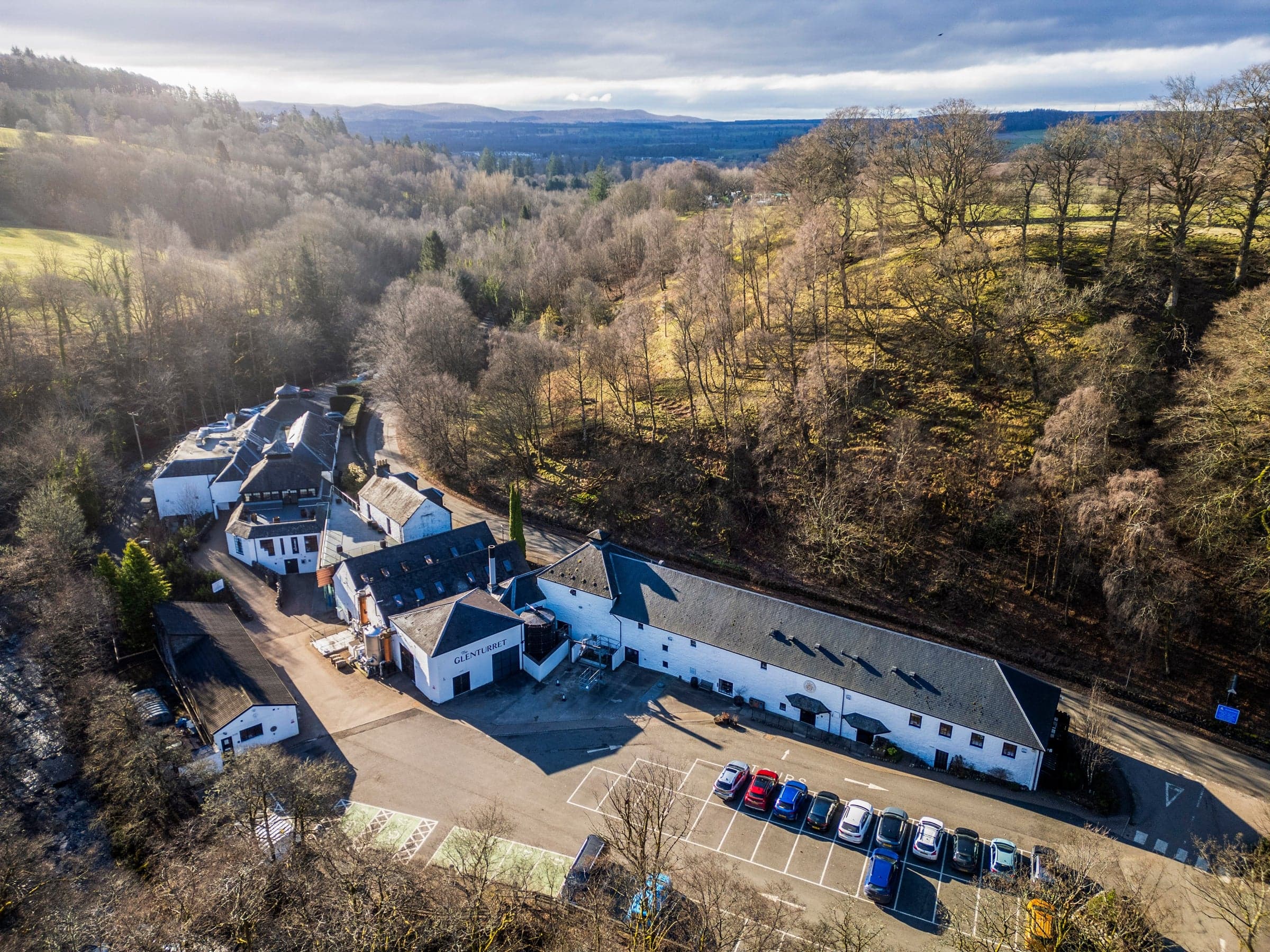 A drone view of The Glenturret distillery in winter — a cluster of low whitewashed buildings with grey slate roofs strung along the road in the wooded Strathearn glen, the Ochil Hills in soft cloud beyond.