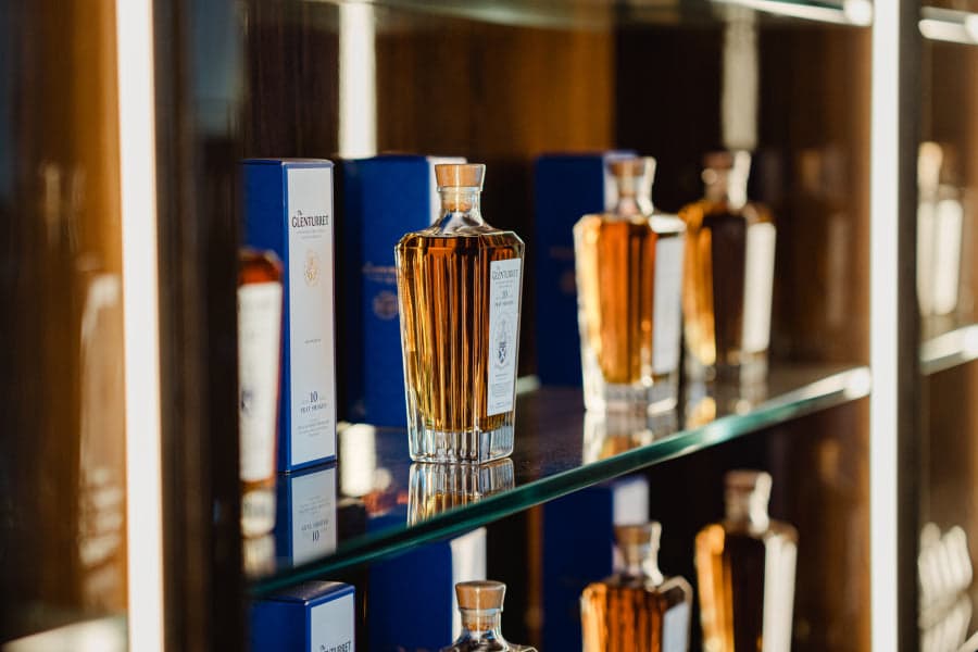 A close shot of three fluted Glenturret crystal decanters on a glass shelf — a pale single malt in the centre bottle catching the light, navy blue Glenturret presentation boxes ranged behind, the warm interior of the Lalique Bar reflected in the mirror behind.