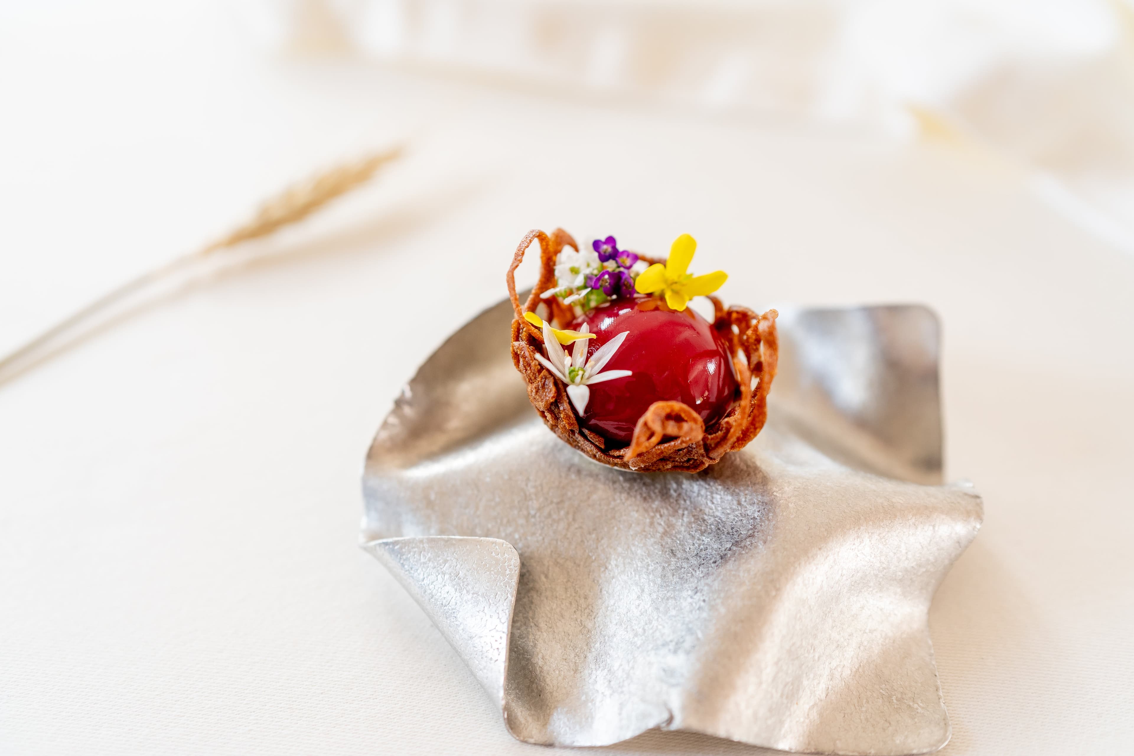 A small petit four — a glossy red sphere of raspberry liver pâté in a fried lace tuile basket, finished with three white starflowers and a yellow violet — set on a hand-pressed silver foil sculpture on a cream tablecloth, a single dried wheat ear soft in the foreground.