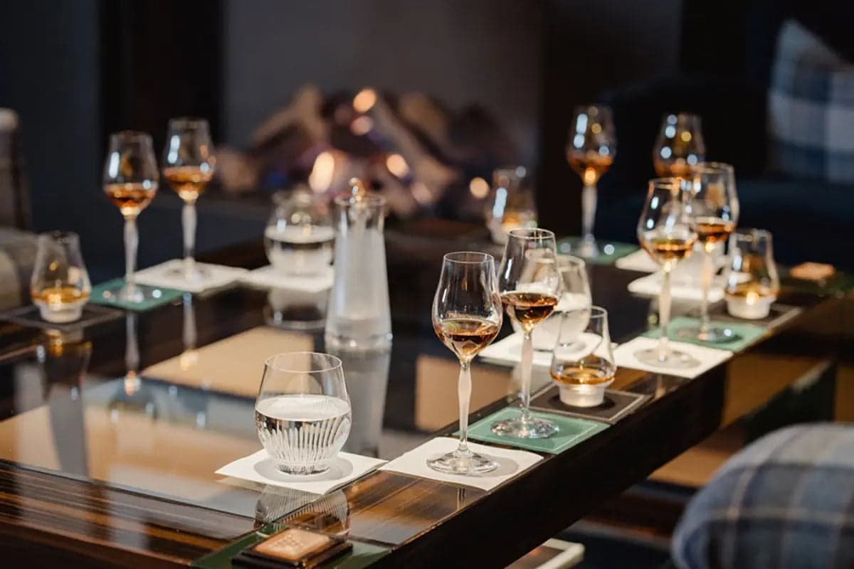 A long whisky tasting table at the Lalique Bar — eight Glencairn copita glasses on green cards in a line down a polished dark wood table, each holding a different Glenturret expression, a low log fire blurred behind in the dark room.