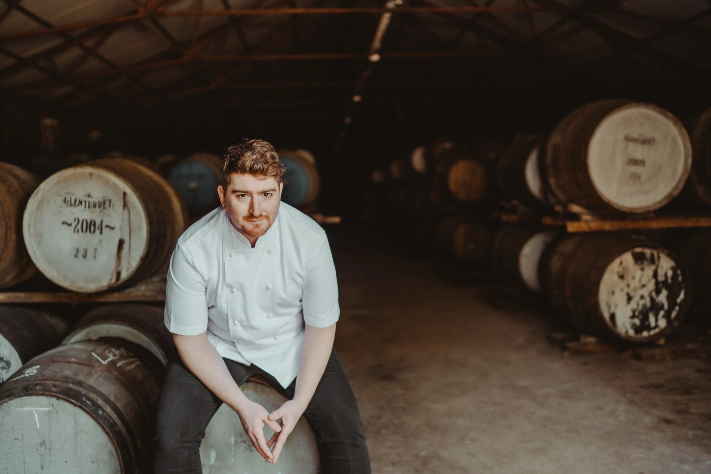 Mark Donald in a short-sleeved white chef's jacket sat on the head of a Glenturret cask in the dunnage warehouse — rows of stencilled oak whisky barrels on either side dark in the half-light, his hands folded on his knee.