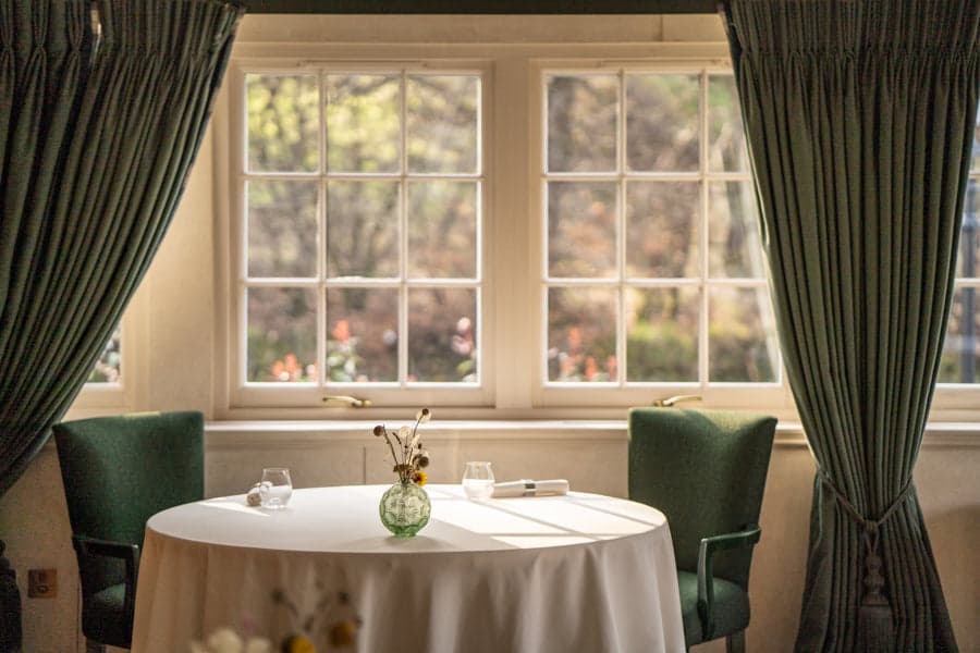 A round two-top at The Glenturret Lalique dressed in white linen with a small green pressed-glass bud vase, set between two fluted dark green wing-back chairs in front of a sash window onto autumn woodland with green velvet curtains drawn back at either side.