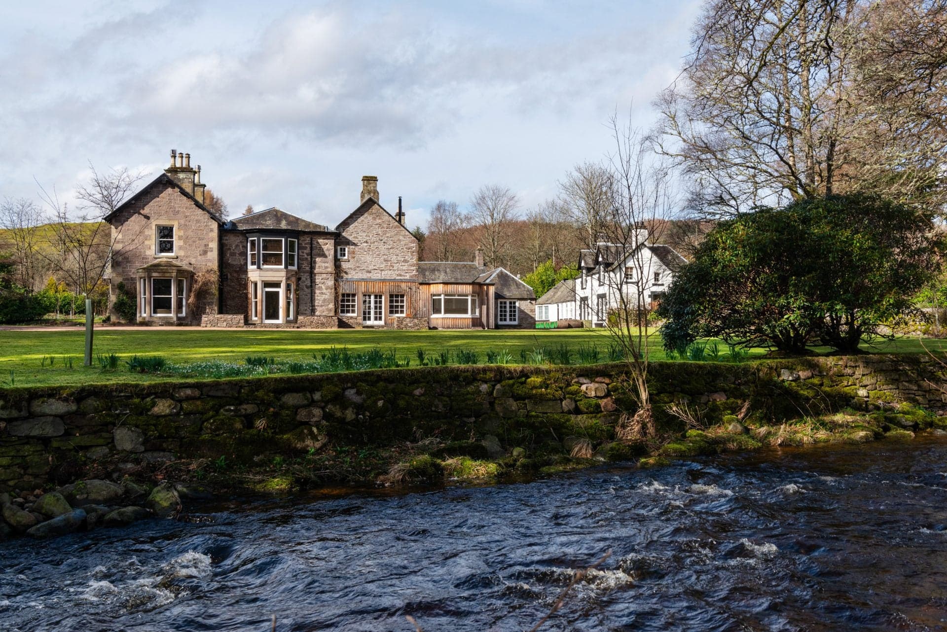 The Aberturret Estate House — a pale-stone Georgian Scottish house with two-storey bay windows and a smaller Victorian wing, set on a manicured lawn behind a low stone wall, the River Turret running fast over rocks in the foreground and bare trees on the hill behind.