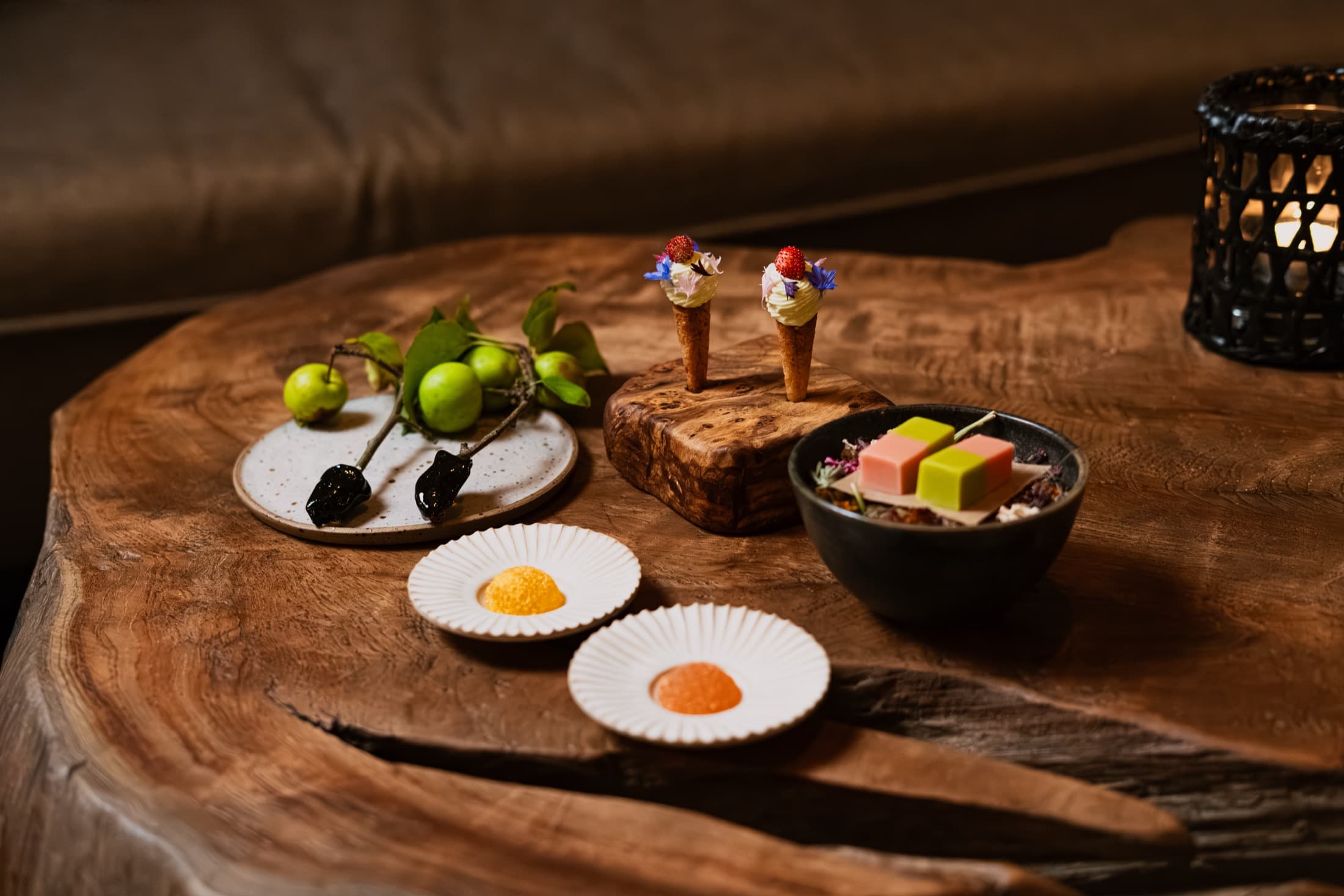 A board of opening petits-fours on a live-edge wooden table in low candlelight — a sprig of green crab apples with two charred fruits, two miniature waffle cones piped with cream and topped with wild strawberries, jellies and pâtes de fruits arranged on fluted white plates.