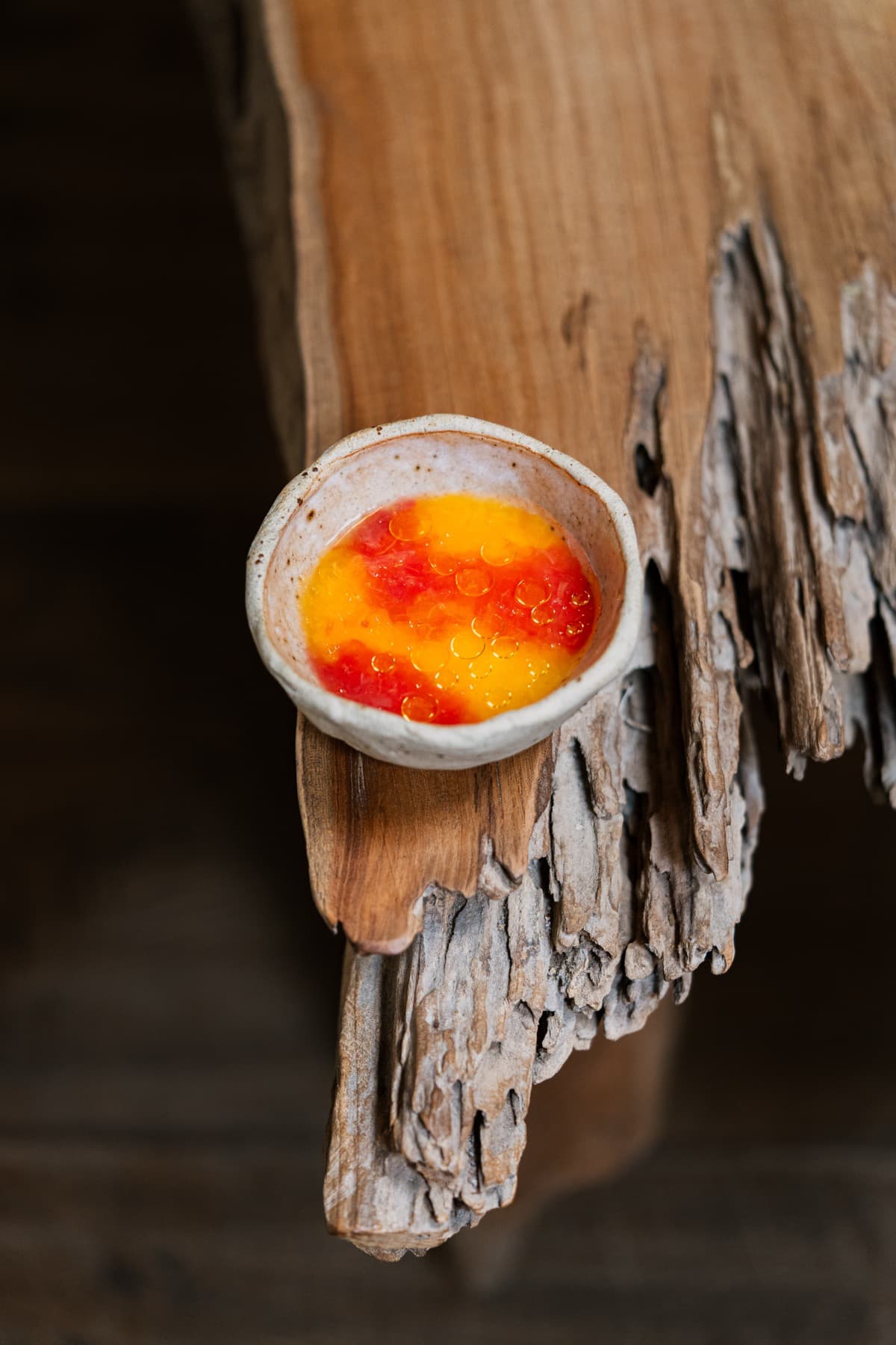 A small hand-thrown ceramic dish balanced on a piece of weathered driftwood — the dish holds a glossy yellow custard topped with diced red strawberry and amber droplets of fruit gel.