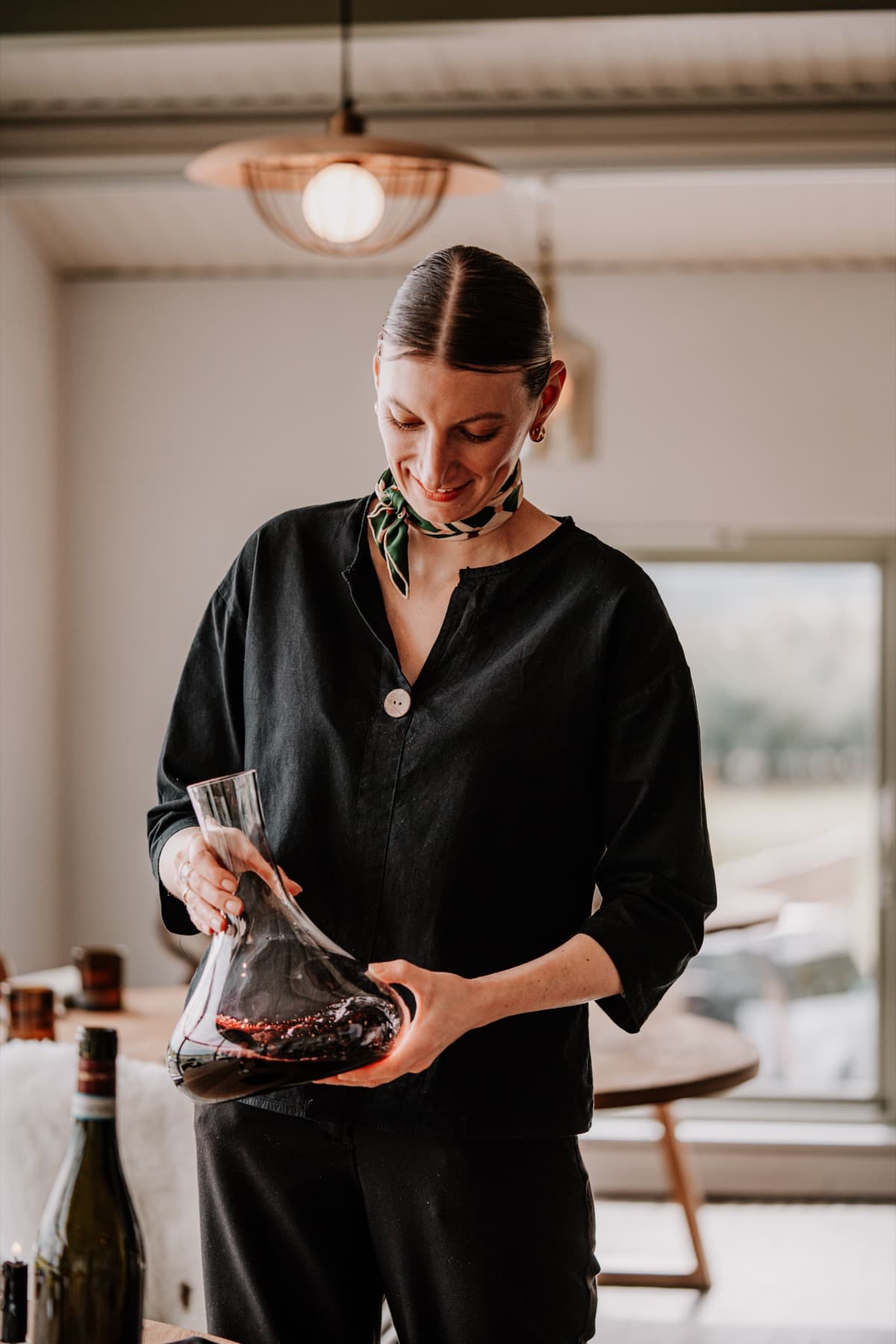 Head sommelier Vanessa Stoltz decanting a red wine in the dining room — black linen blouse, green silk scarf, a copper pendant lamp warm above her, the round oak tables of the room in soft focus behind.