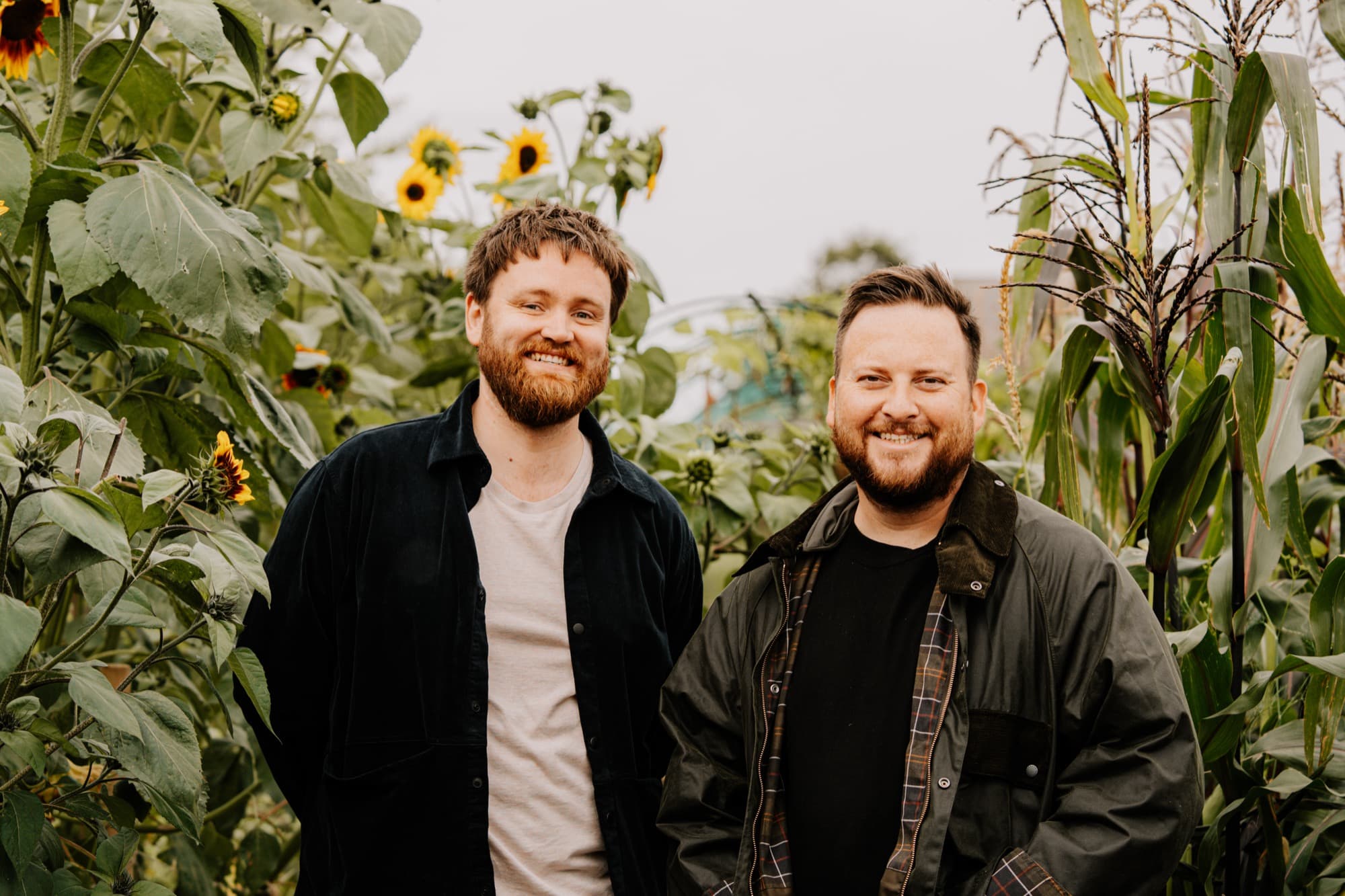 Cal Byerley and Ian Waller standing among rows of sunflowers and sweetcorn in the Vallum Farm kitchen garden — both bearded, in dark workwear and Barbour, smiling into the lens under a flat Northumberland sky.