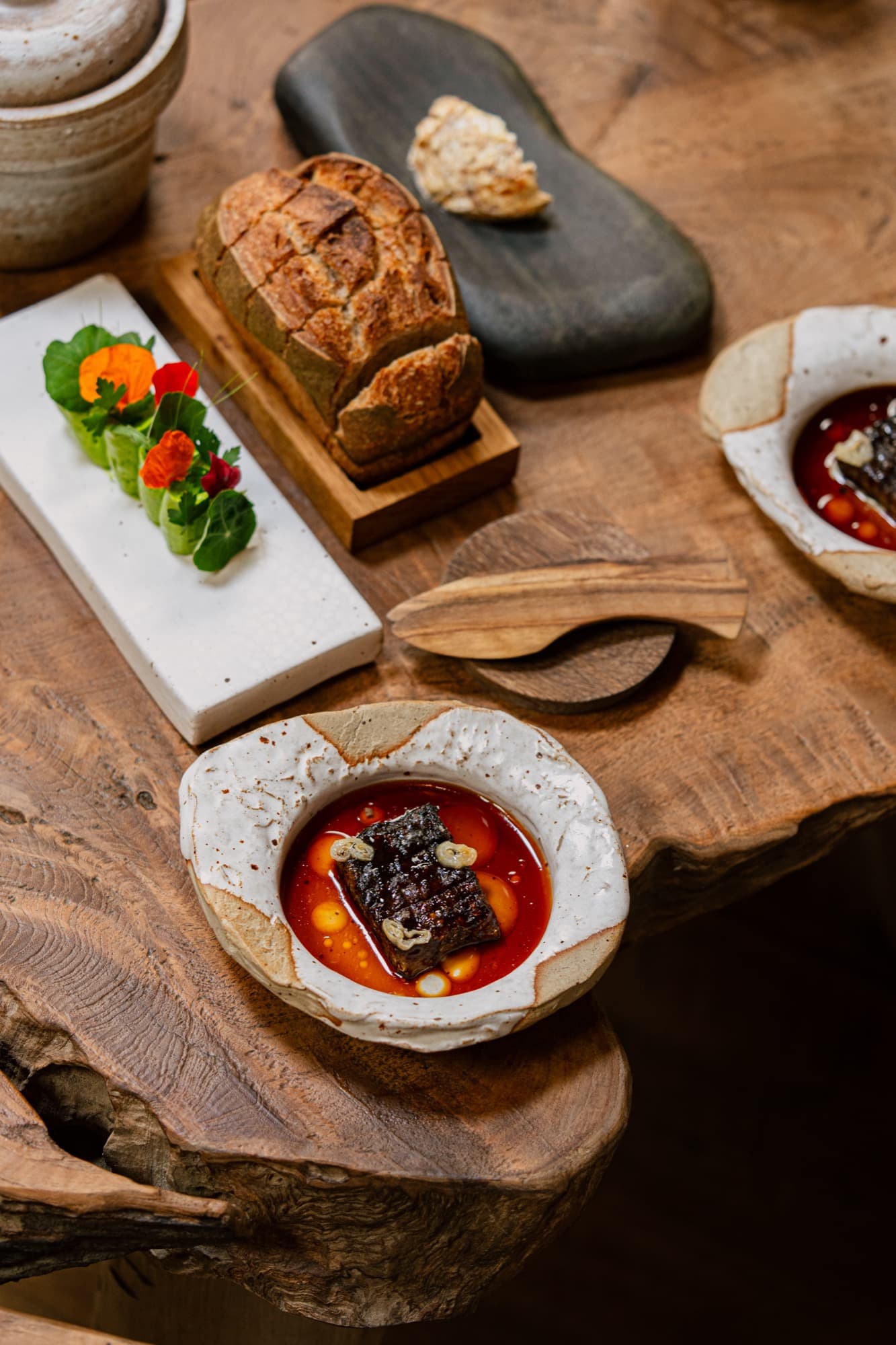 A live-edge oak table laid for an opening course — a small loaf in a cradle, a stone slab with marigolds and salad leaves, and a pale ceramic bowl holding a wood-grilled rib in a deep tomato broth with white beans.