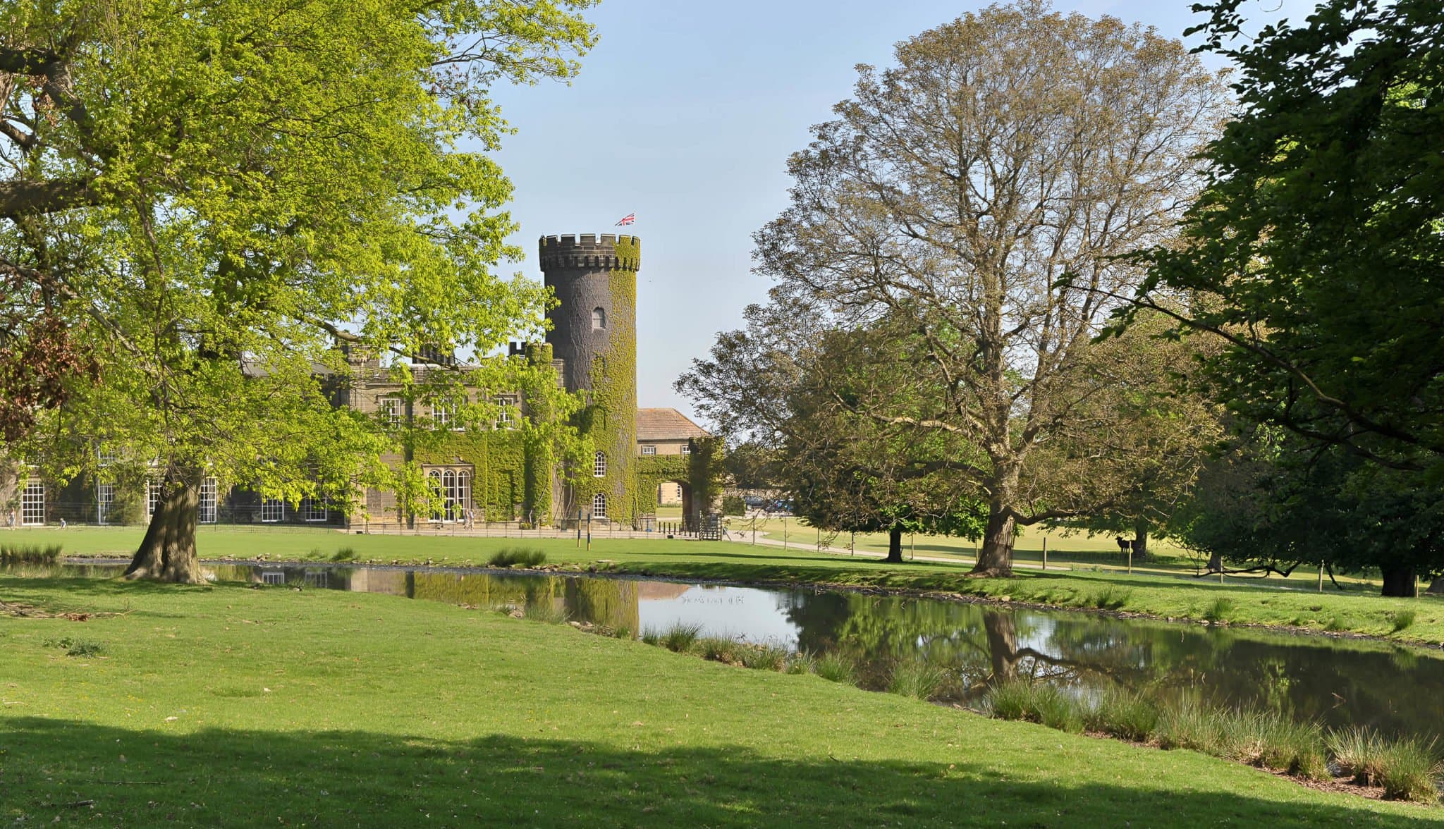 The castellated grey-stone façade of Swinton Park reflected across its lake at midday.