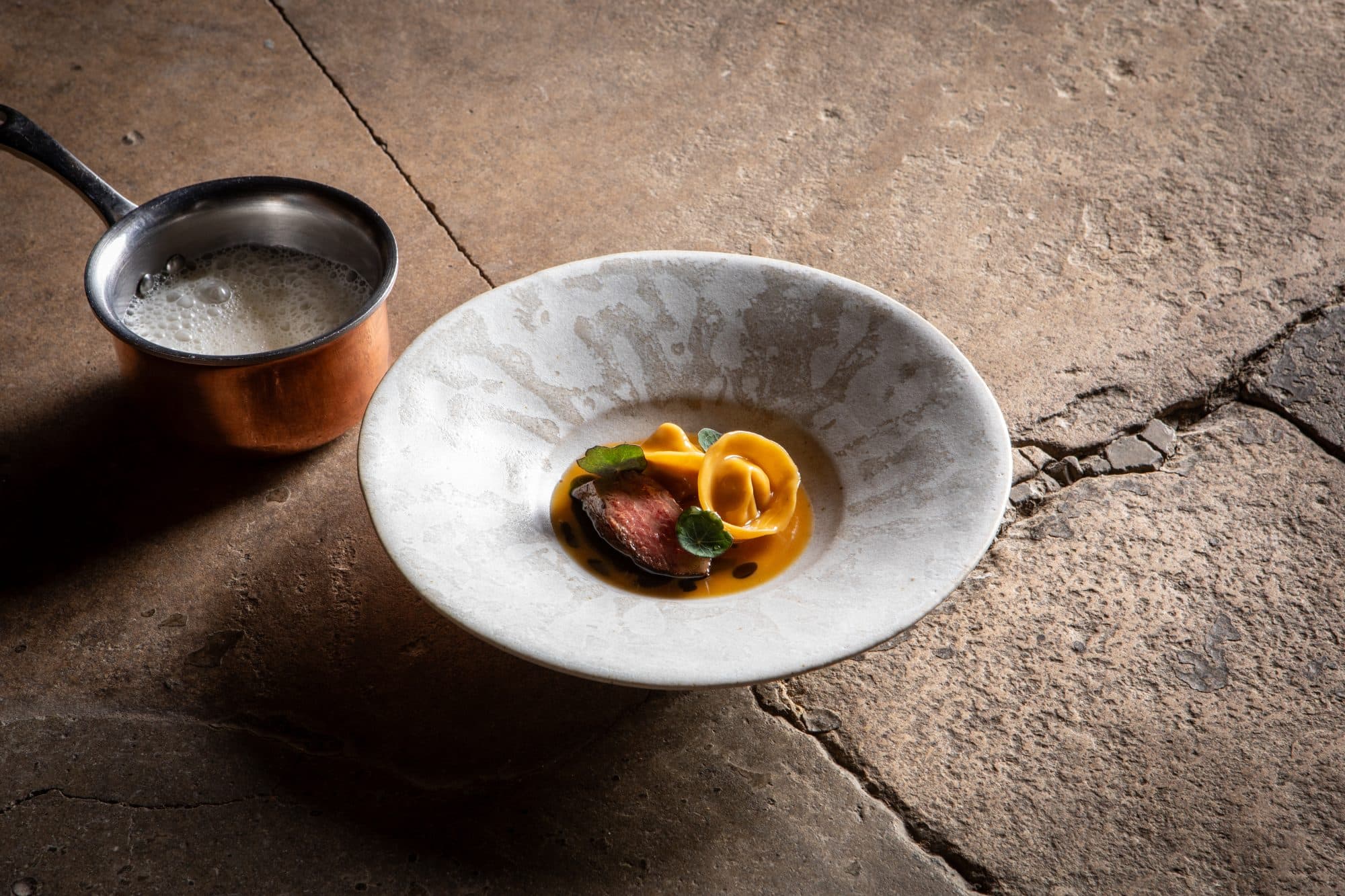 A pale stone-glazed bowl on flagstone — a slice of rosy aged beef in a pool of orange squash purée with a small green herb, a copper saucepan of foamed butter sauce alongside.