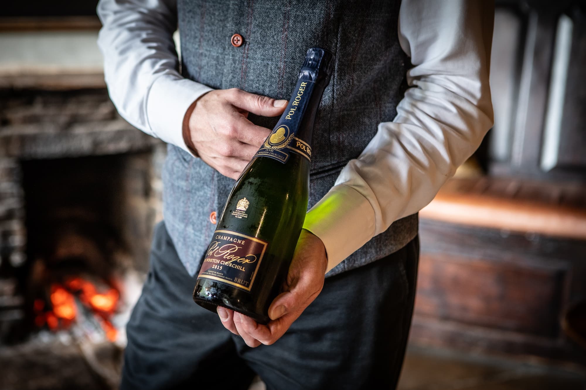 A sommelier in a tweed waistcoat holding a bottle of Pol Roger Sir Winston Churchill 2015 in front of a low fire — close on the hands, the firelight warm in the brick.