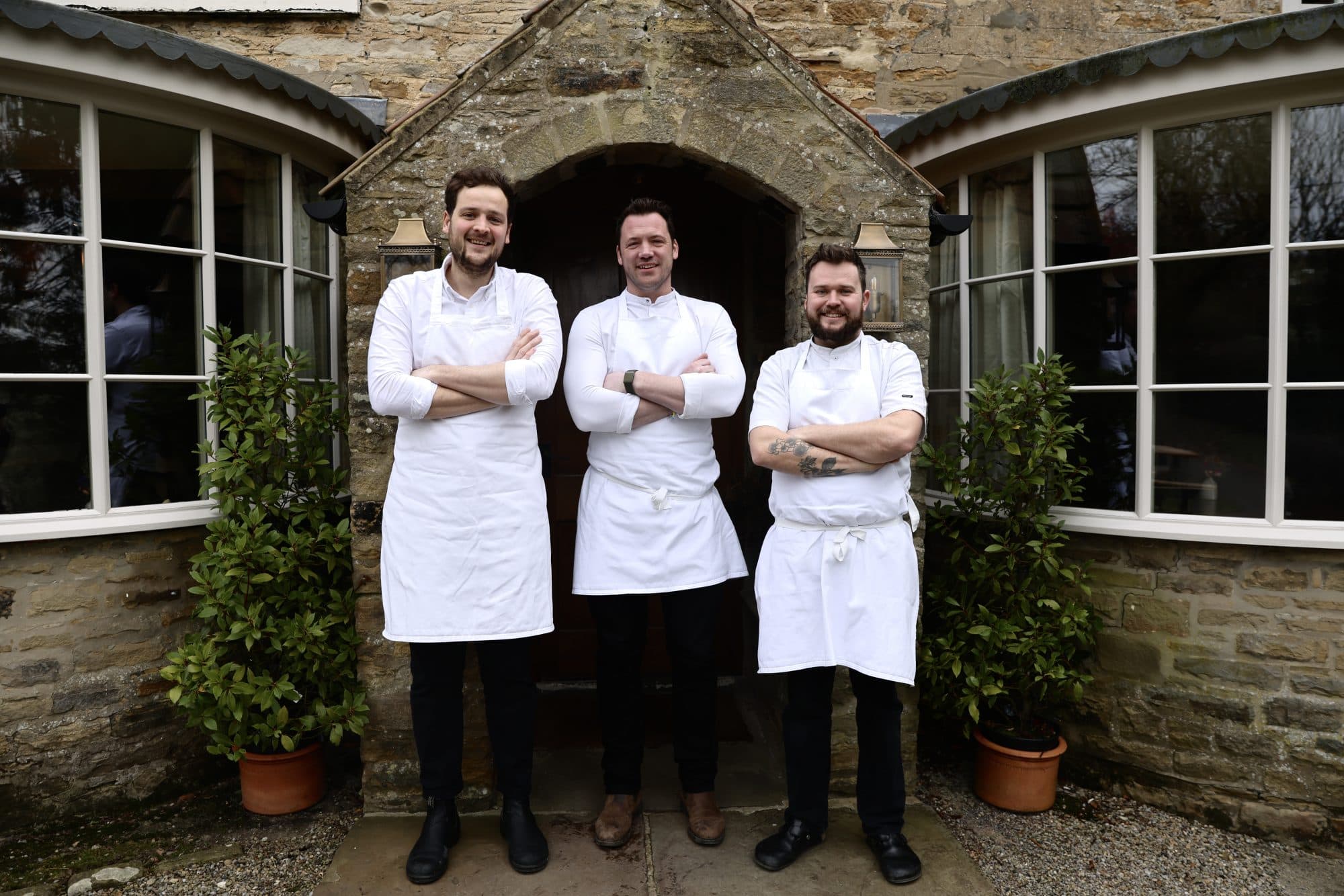 Three chefs in white aprons standing under the stone porch of The Black Swan — Tommy Banks at the centre, head chef and executive chef on either side, the bay windows of the dining room behind.