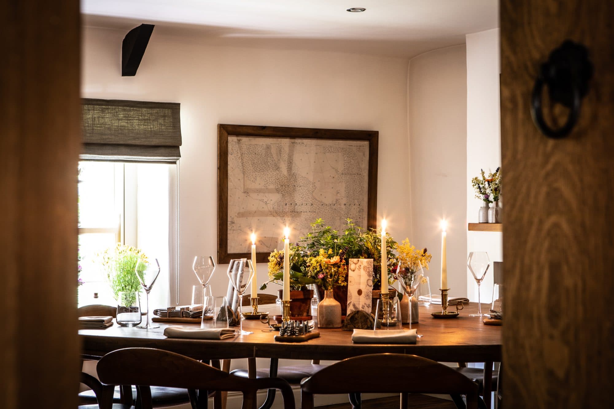 A long oak table laid for service — wildflowers in glass vases, a row of brass candlesticks lit, a framed estate map of Oldstead on the white wall above, the dining room glimpsed through an open oak door.