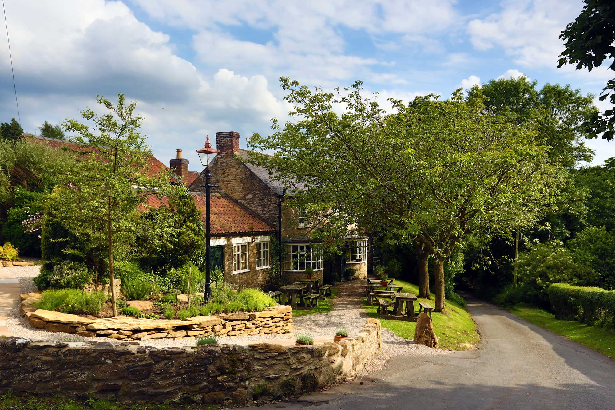 The Black Swan from the village lane — pale-stone pub with red pantiles half-hidden by summer trees, low stone walls and picnic benches at the front, the Yorkshire sky high and bright above.