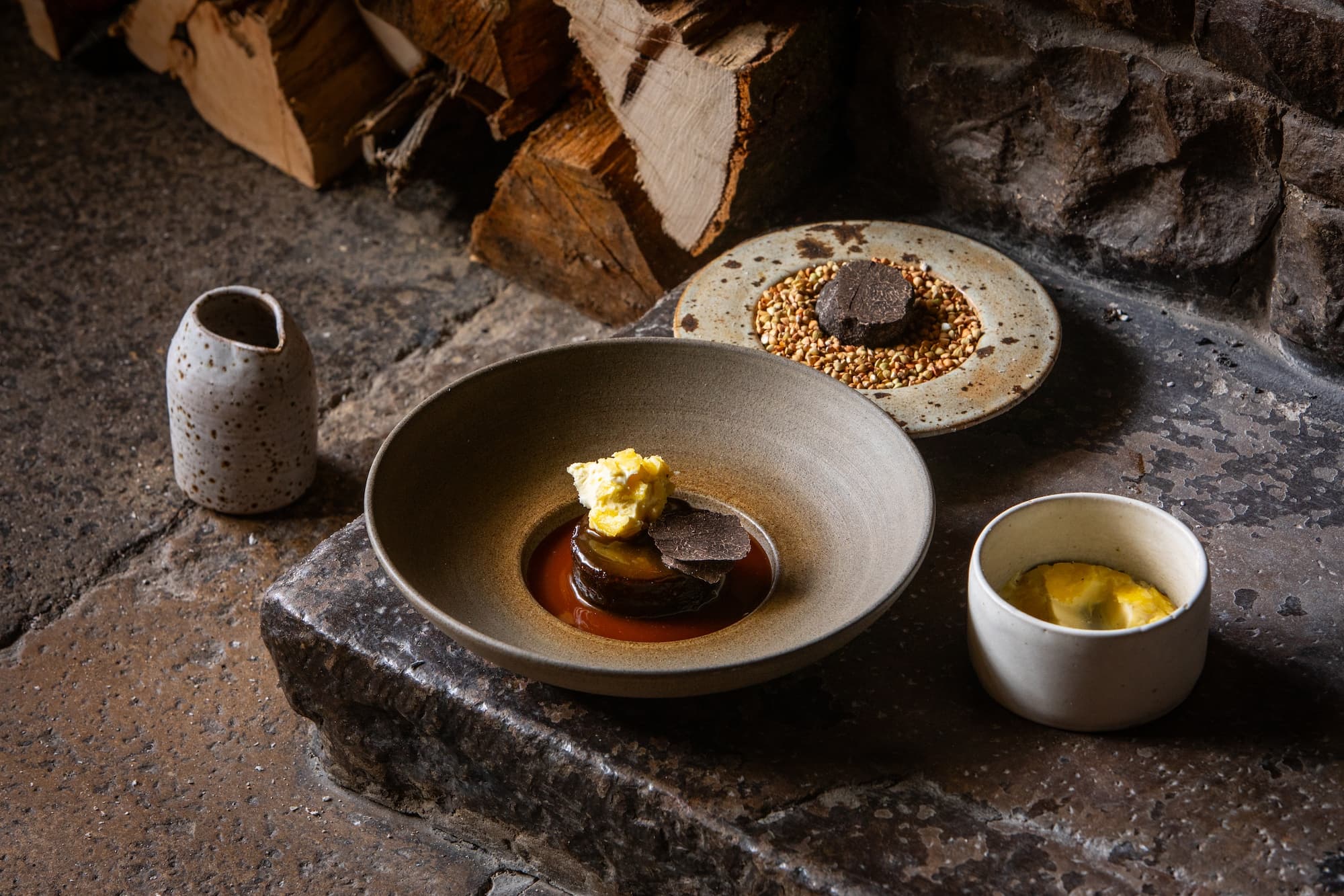 A dark-glazed bowl on a stone hearth — a chocolate and beetroot dessert with a quenelle of cream, a small ramekin of yellow custard alongside, and a side plate of granola set against a stack of split logs.