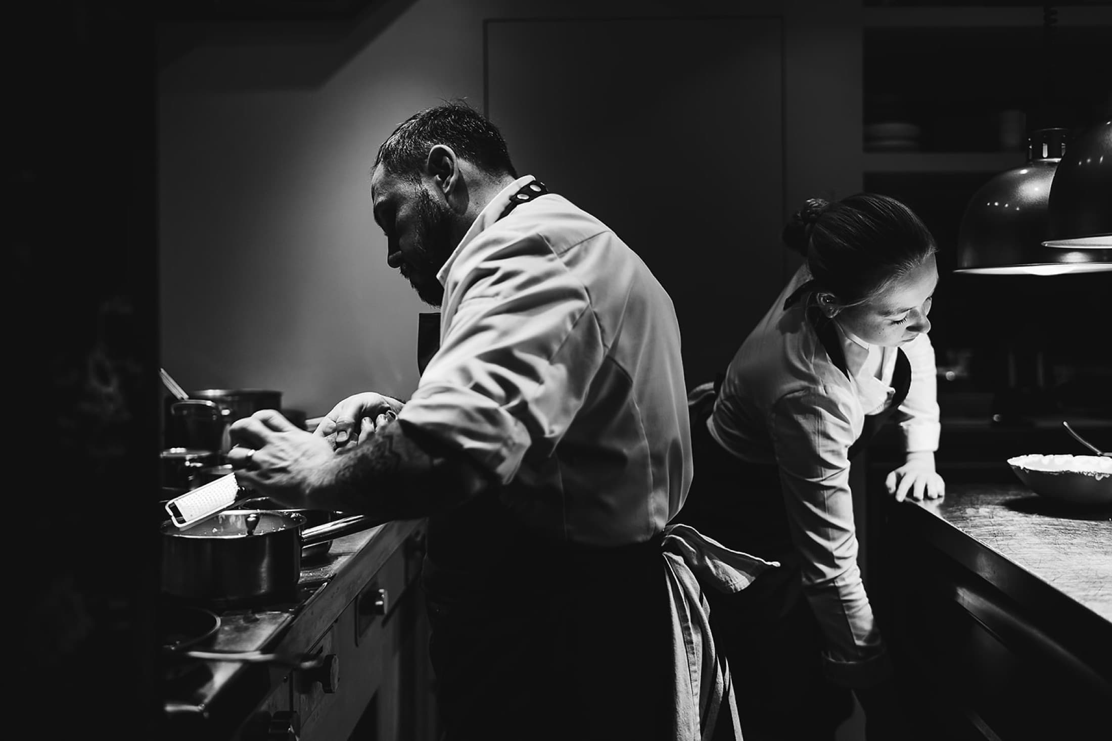 A black wall in the dining room covered in chalk sketches of dishes — chive-oil dressing diagrams, a cherry sorbet plot, slow-cooked herb notes — the writing softened by warm bokeh from a candle in the foreground.