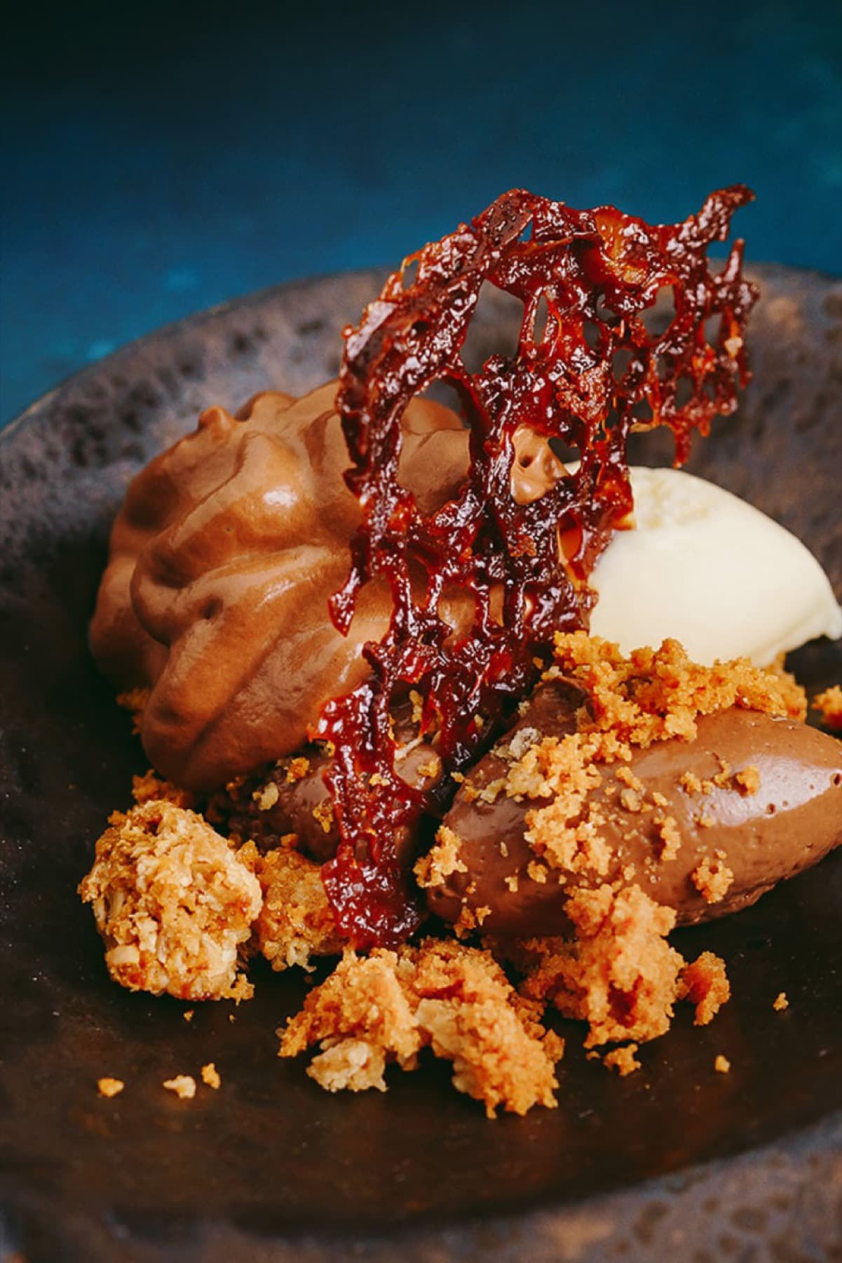 A dark stoneware bowl set on a near-black surface — a quenelle of bitter-chocolate mousse, a quenelle of paler chocolate ice cream, a fragile lattice of dark caramel set on top, oat crumb scattered across the plate.