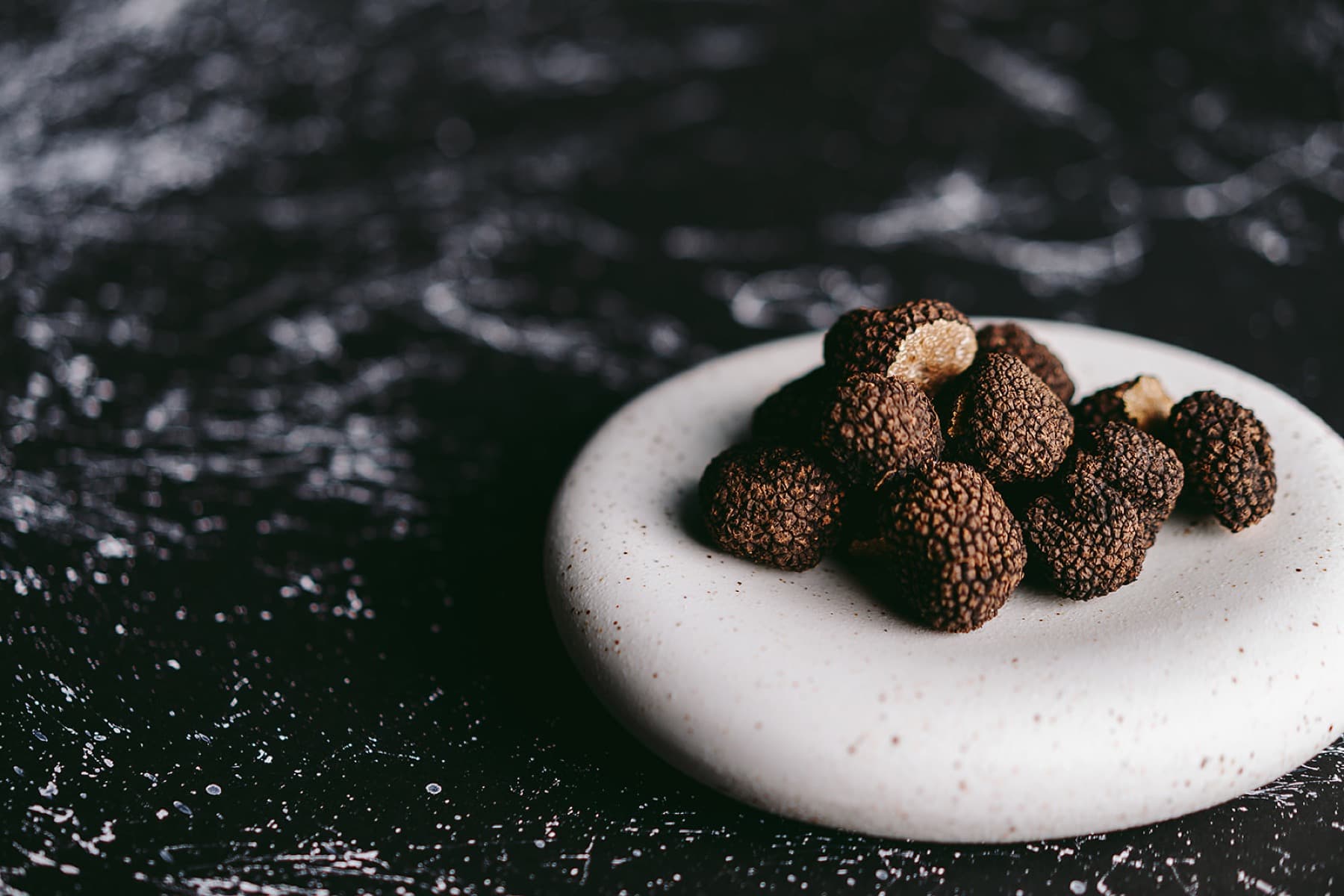 A small white speckled ceramic dish on a black marbled surface — a careful pile of dried black truffles, the flesh of one cut to expose the cream interior, the rest left whole.