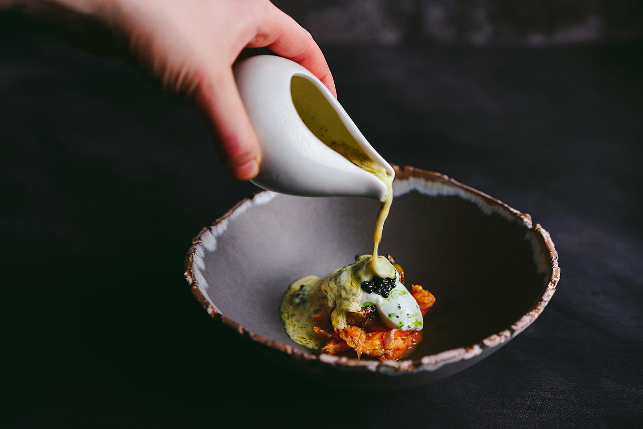 A hand pouring a green herb sauce from a small white jug into a stone bowl — beneath the sauce a glossy crustacean tail with caviar and crème fraîche, a thin lime zest scattered across the dressing.