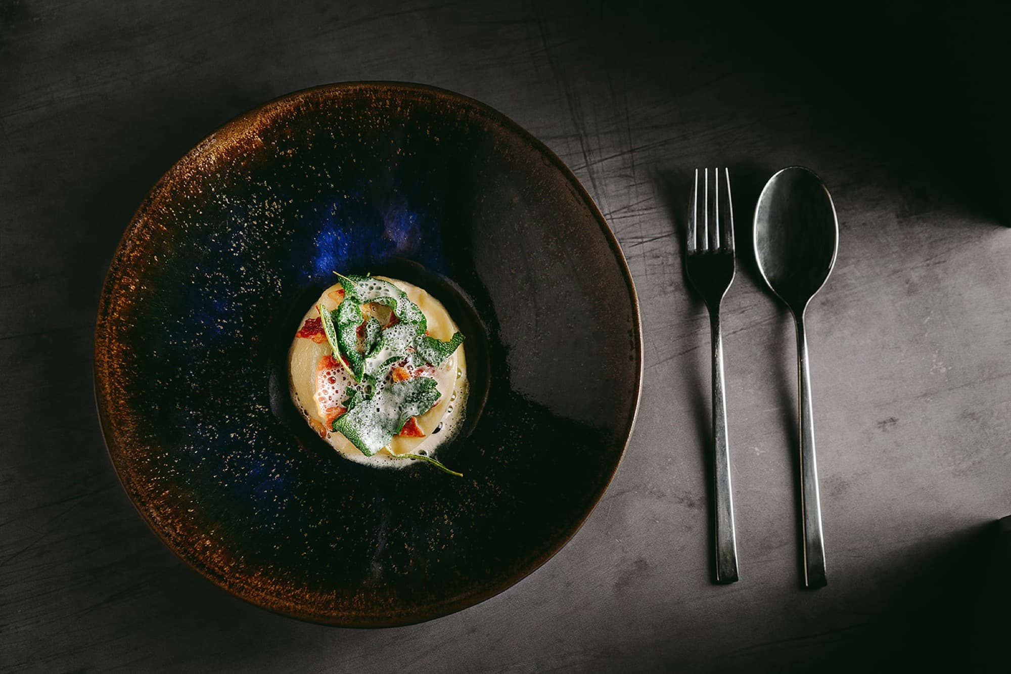 Top-down view of a deep midnight-blue ceramic bowl on a dark slate counter — at its centre a small mound of pale custard, herb leaves, diced chorizo and a fine green-oil foam; a steel spoon and fork laid in line beside it.