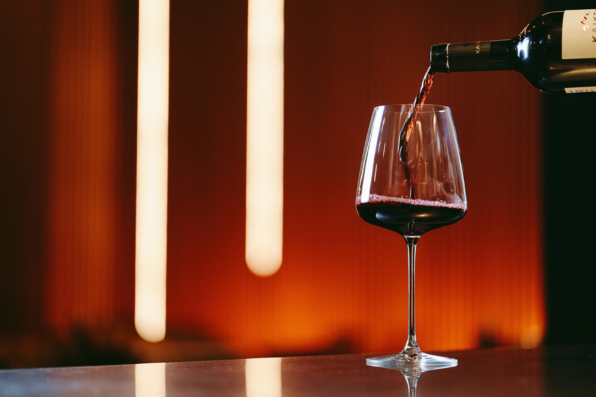 A glass of red wine being poured from a dark bottle in low light — vertical bands of warm orange light glowing on a slatted timber wall behind the glass, the polished bartop reflecting the stem.