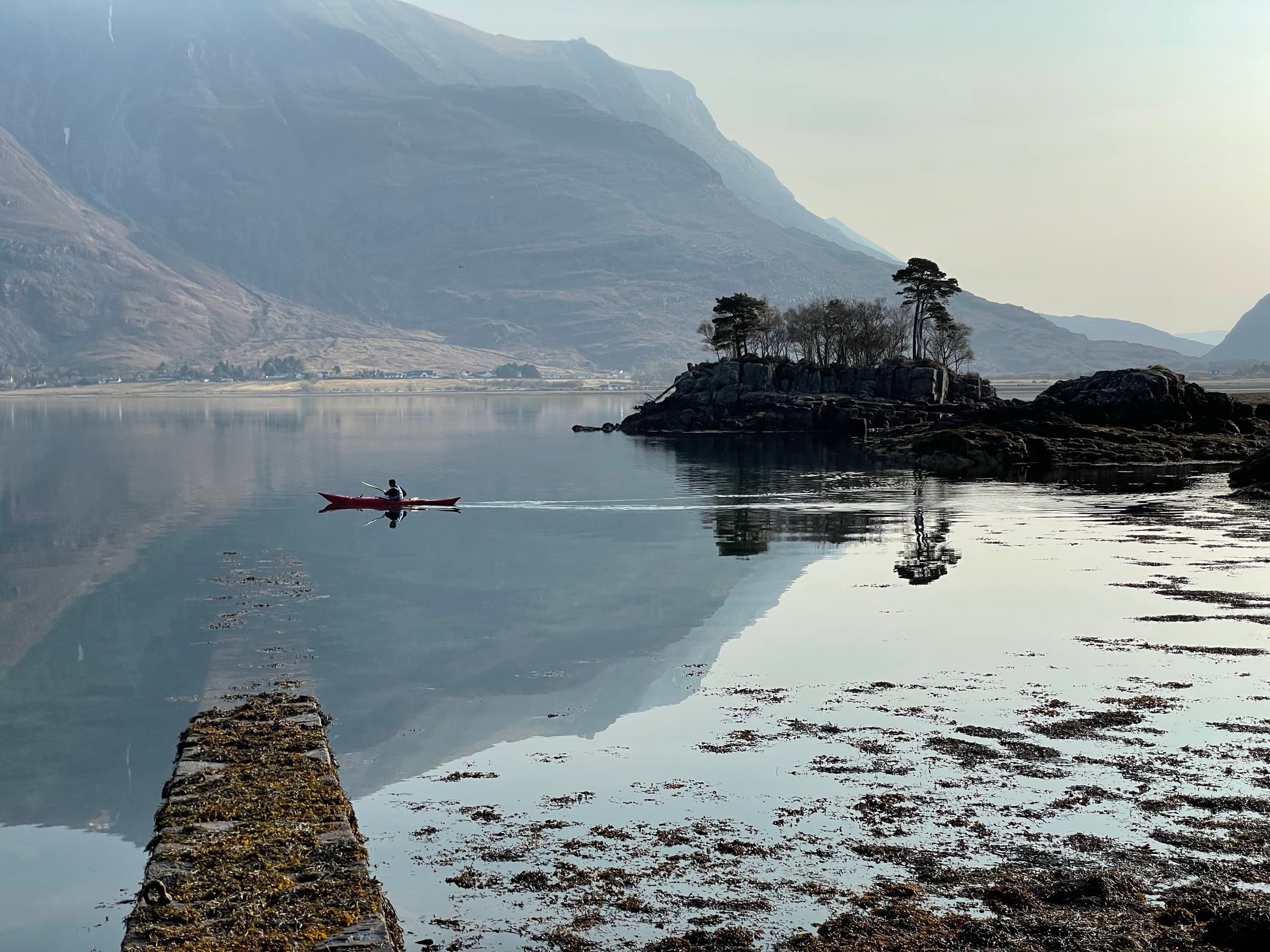 A sea kayaker crossing a glassy stretch of Loch Torridon at dawn, a small island of Scots pines in the middle distance and the mountain beyond it.