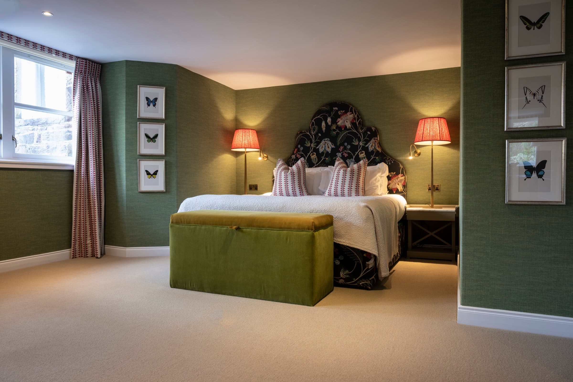 A bedroom in the 1887 Suite — moss-green walls, a floral embroidered headboard, scarlet lampshades and a moss velvet ottoman at the foot of the bed.