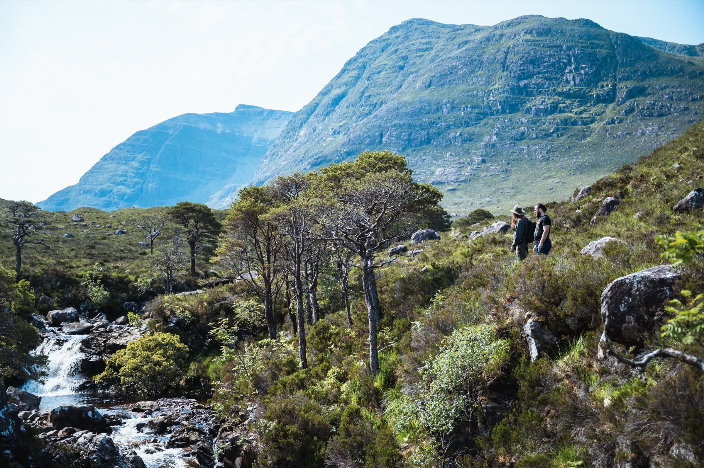 Two walkers crossing a grassy slope above a small waterfall, the long ridge of a Munro filling the sky behind them.